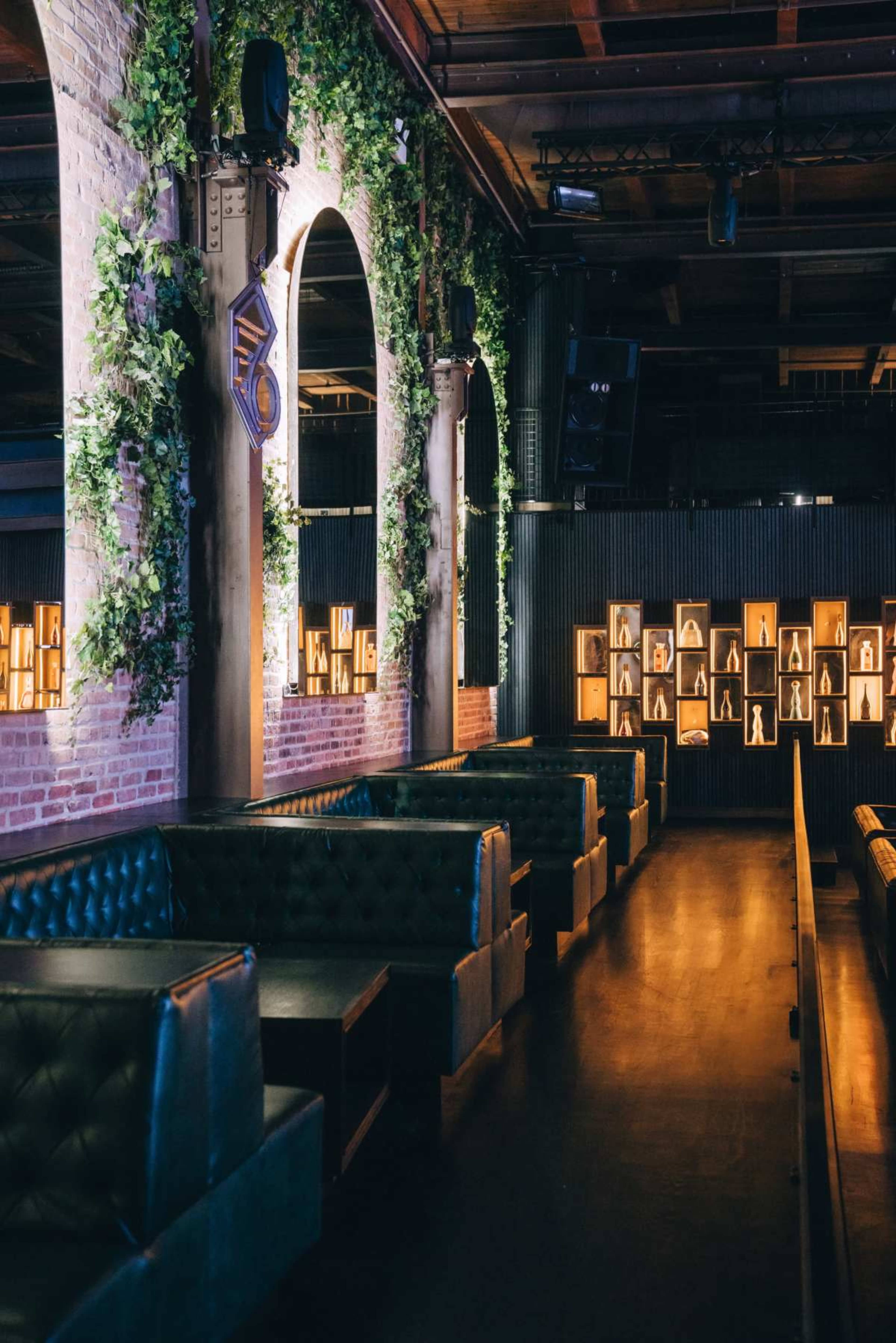The image shows a dimly lit lounge with plush seating, brick walls adorned with greenery, and display shelves featuring framed artworks.