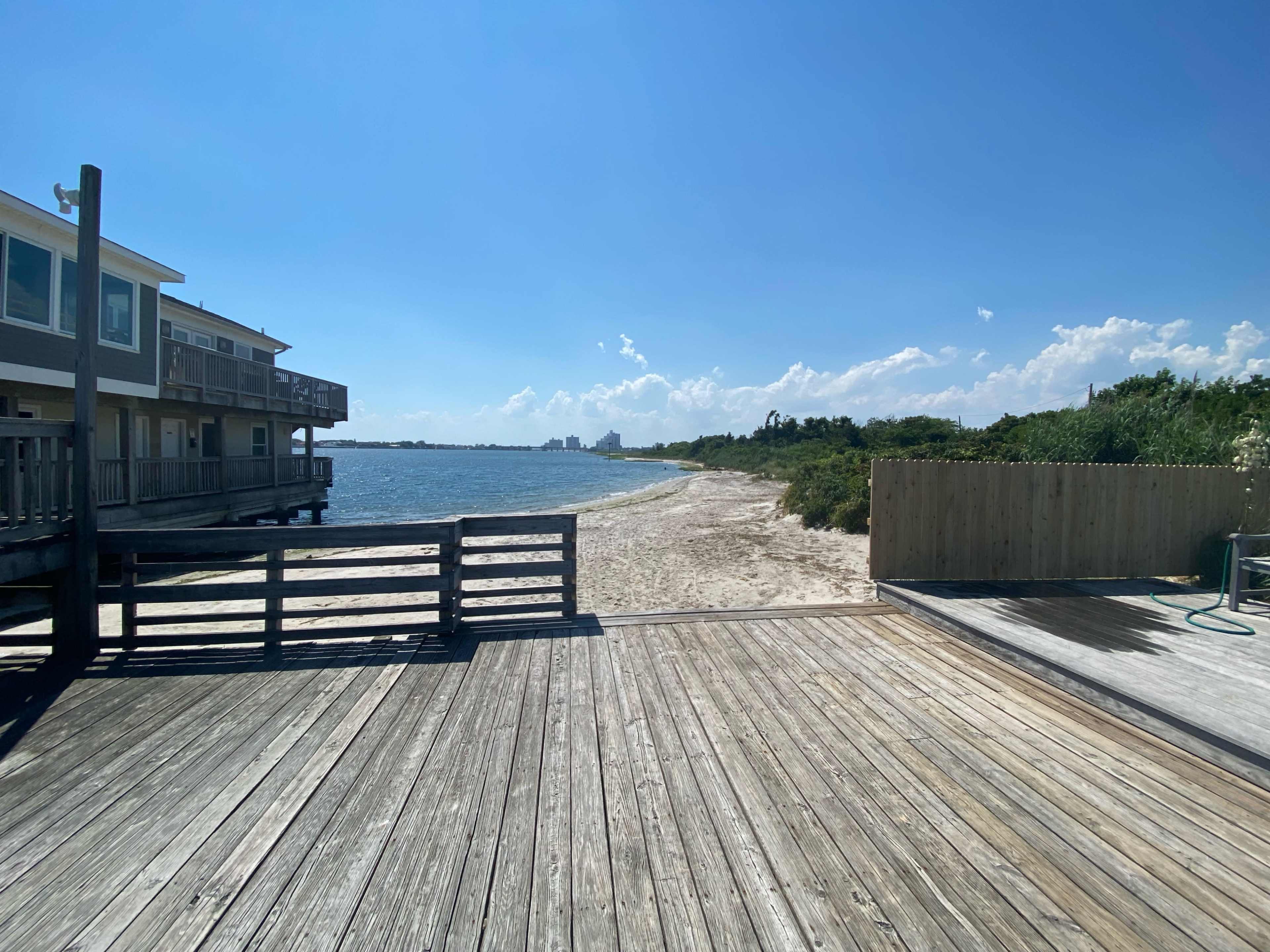 A wooden walkway leads to a sandy beach beside a calm body of water under a bright blue sky.