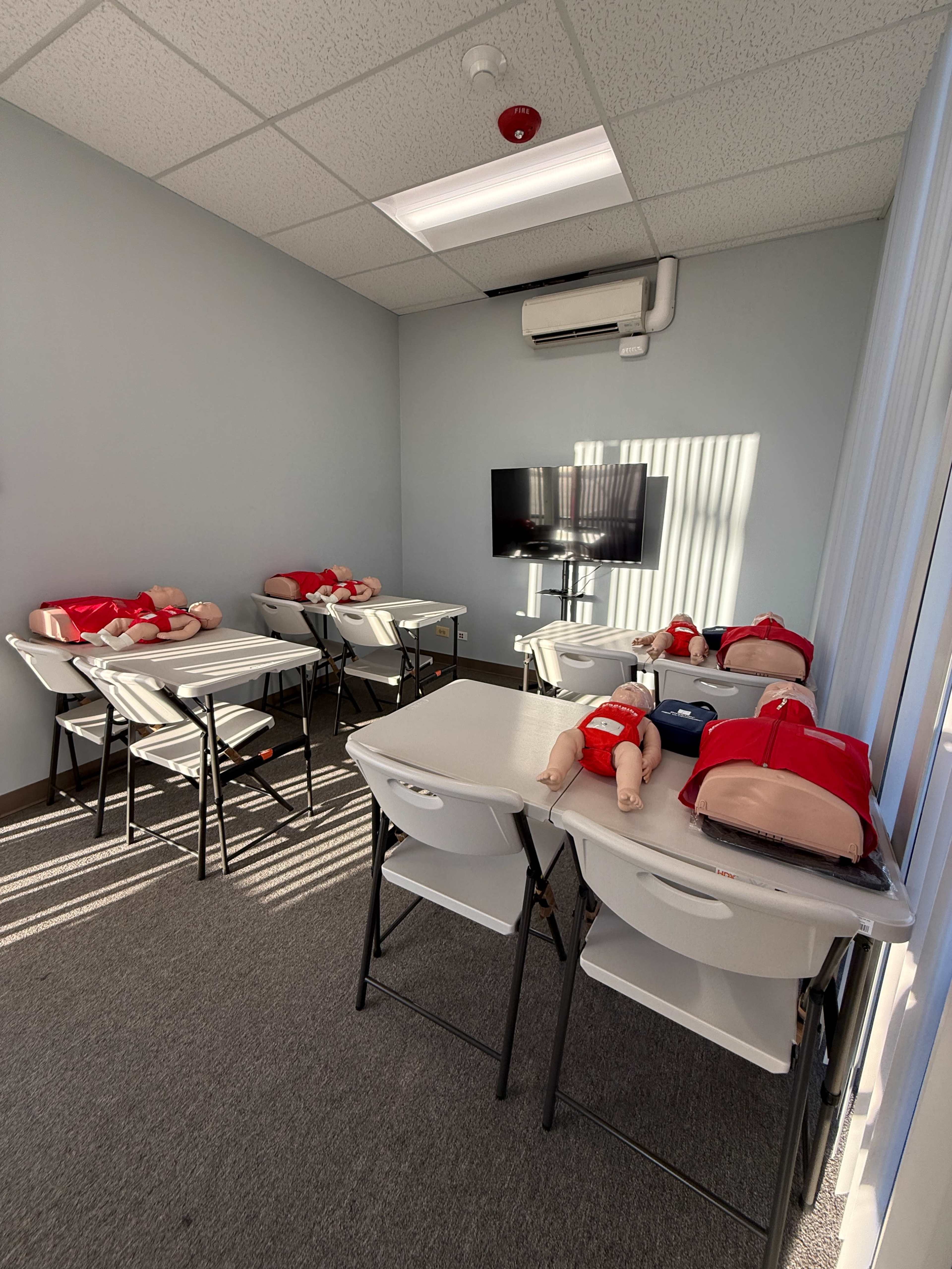 The room contains several folding tables with training mannequins lying on them, and a television mounted on the wall.