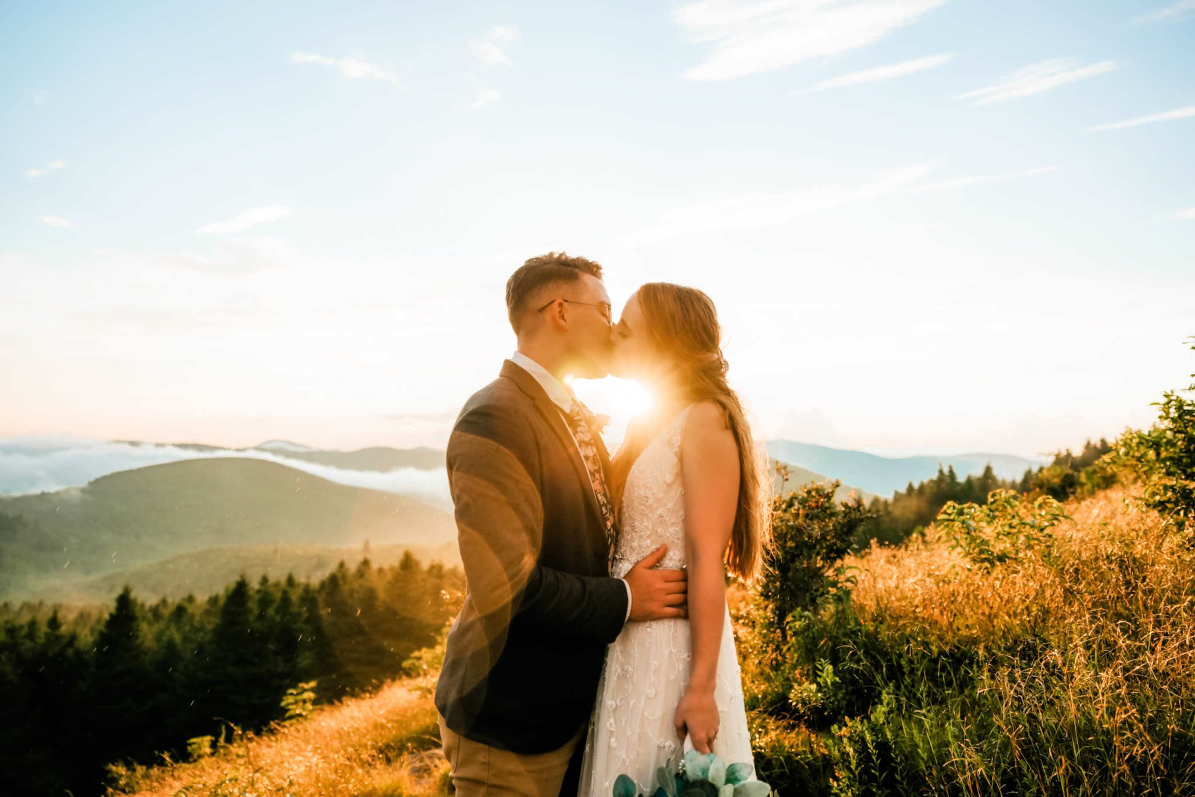 A couple stands in an open field at sunset, embracing and kissing with mountains in the background.