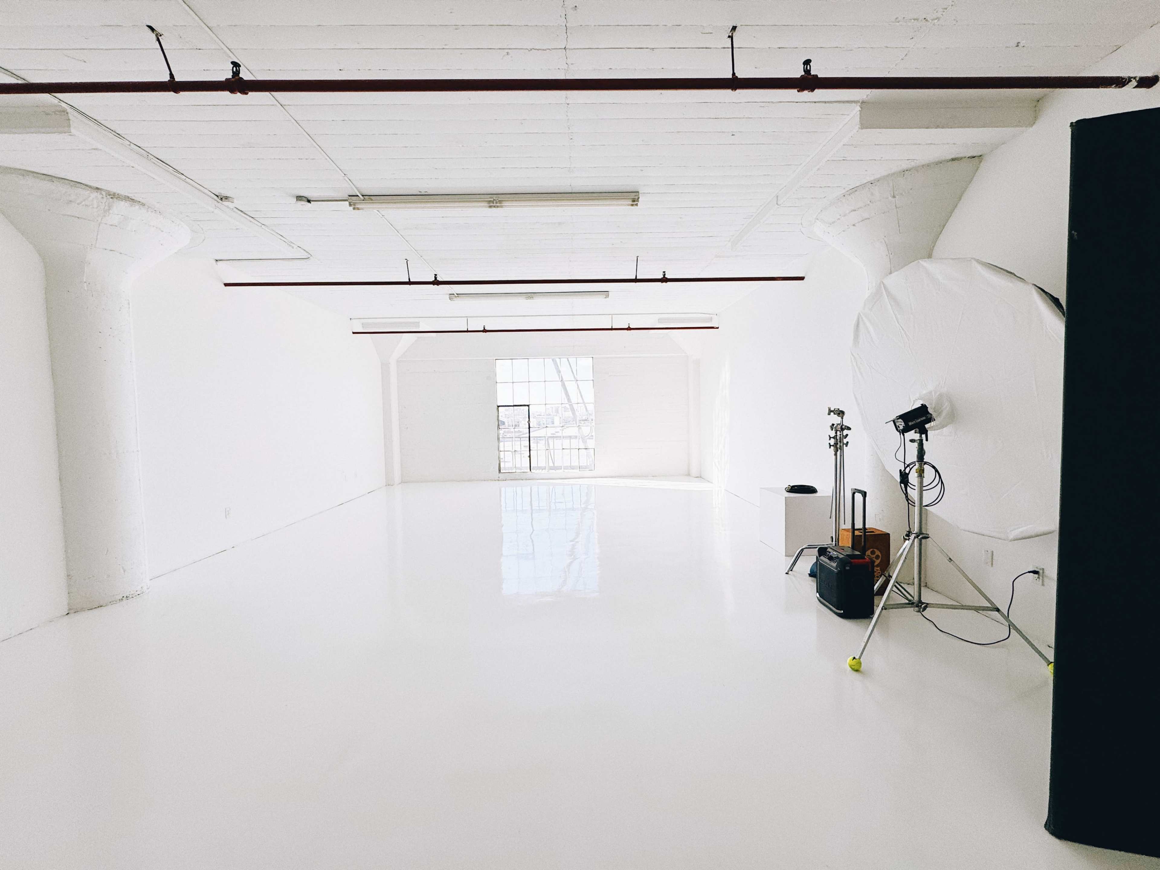 An empty studio space with white walls and a polished floor, featuring a large window and some lighting equipment in the corner.