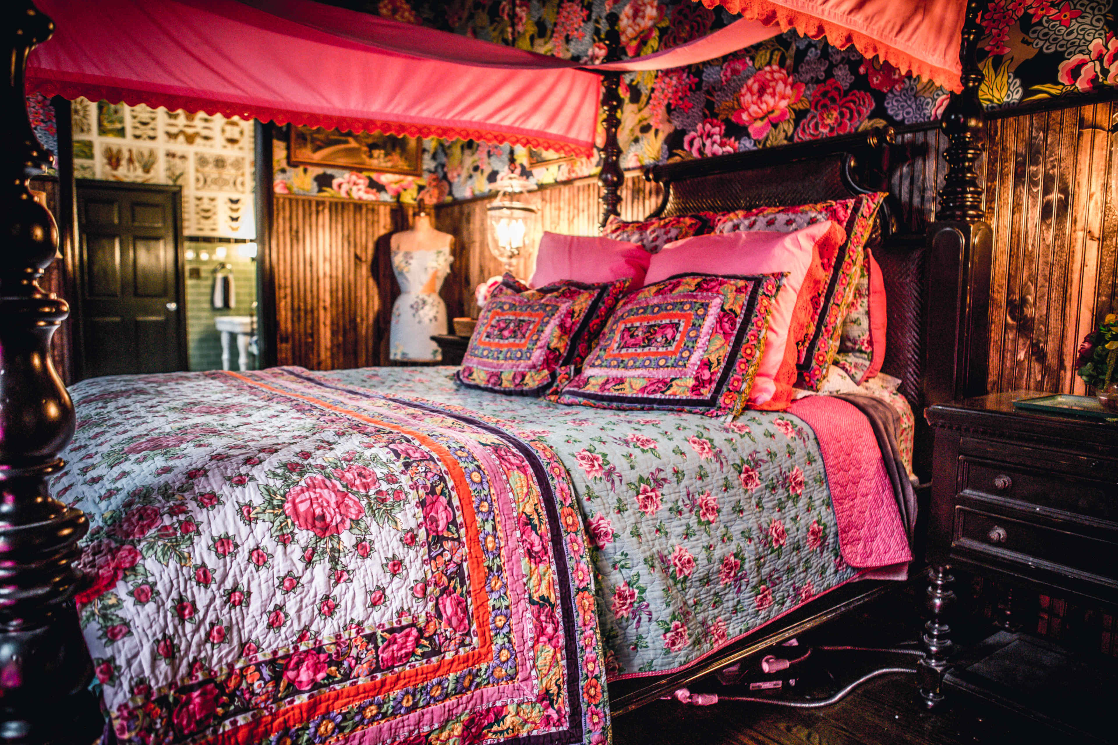 A canopy bed with a floral quilt and matching pillows is set against a vividly patterned wallpaper in a decorated bedroom.