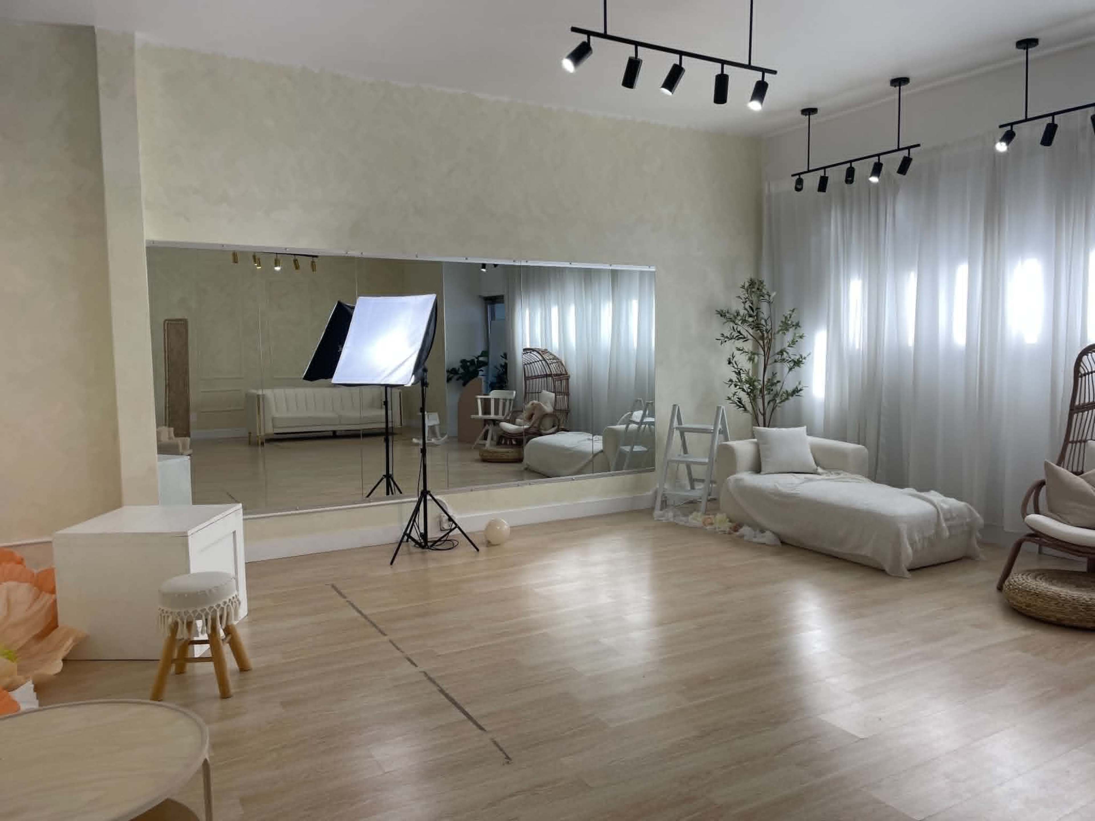 The image shows a spacious, softly lit room with a large mirror, a sofa, a light-colored chair, and studio lighting equipment set up for photography.