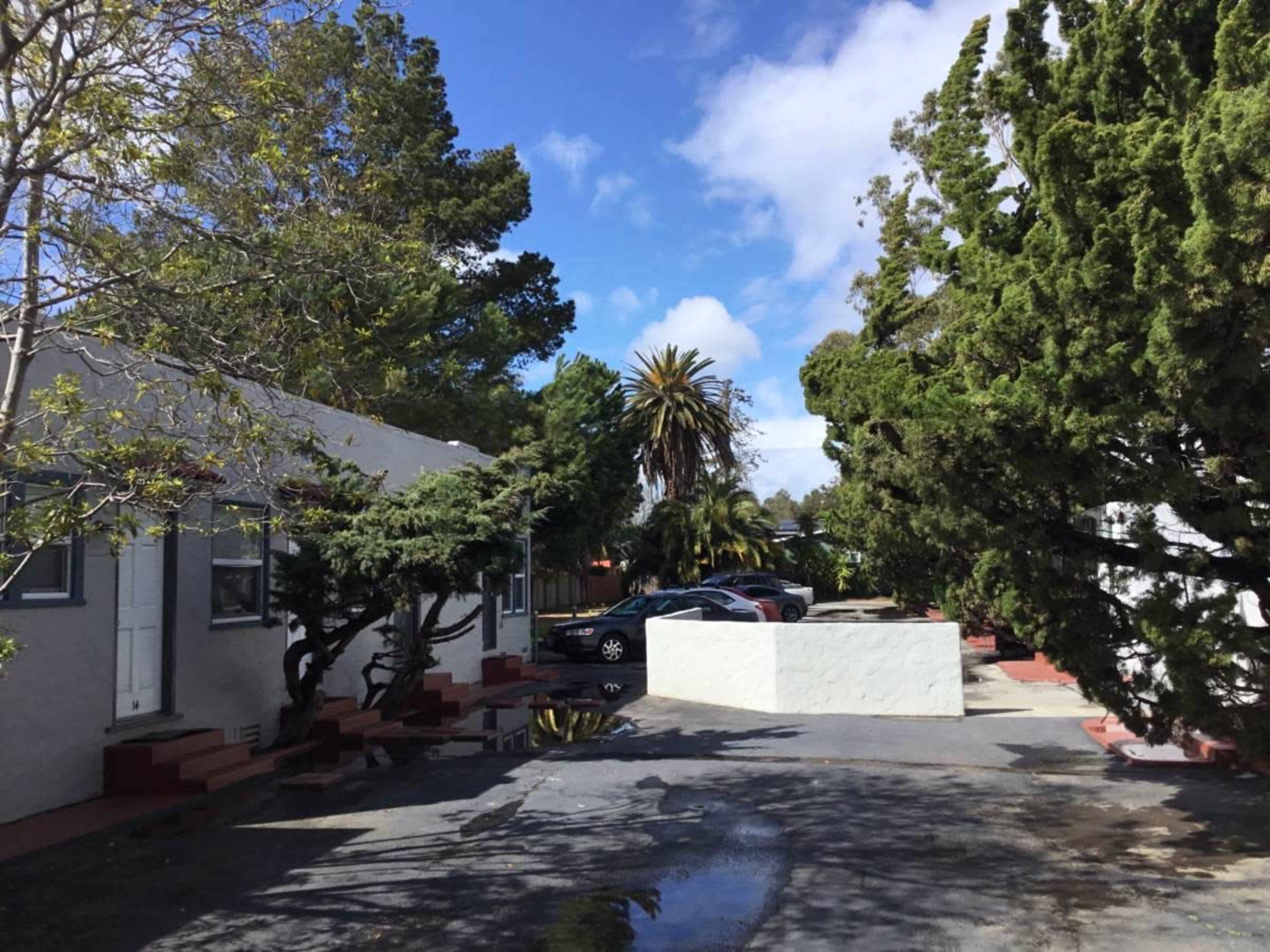 The image shows a pathway lined with trees, leading to a parking area next to a row of single-story buildings under a partly cloudy sky.