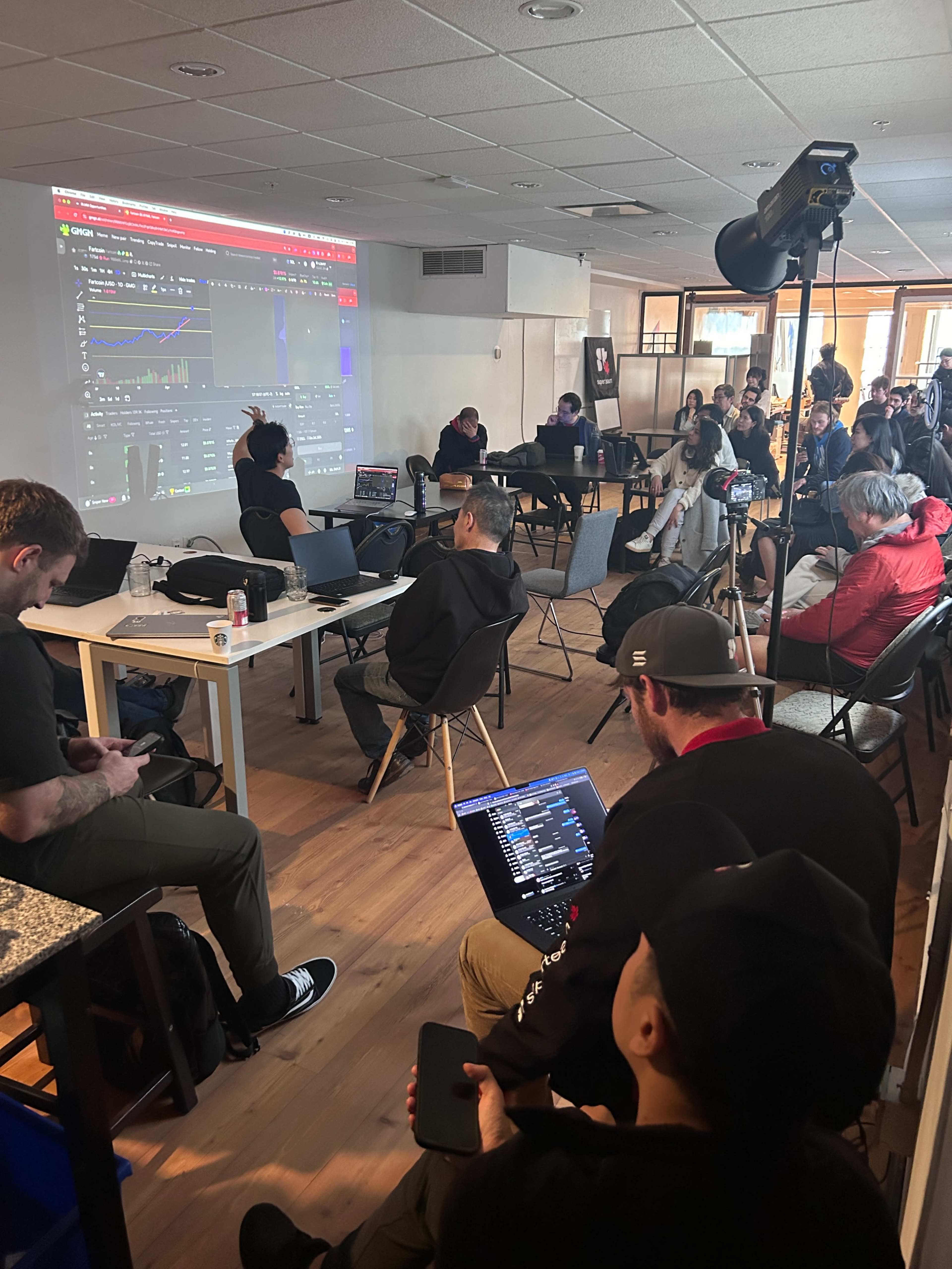 A group of people is seated in a bright room, attentively watching a presentation on a large screen while various electronic devices are visible on tables.