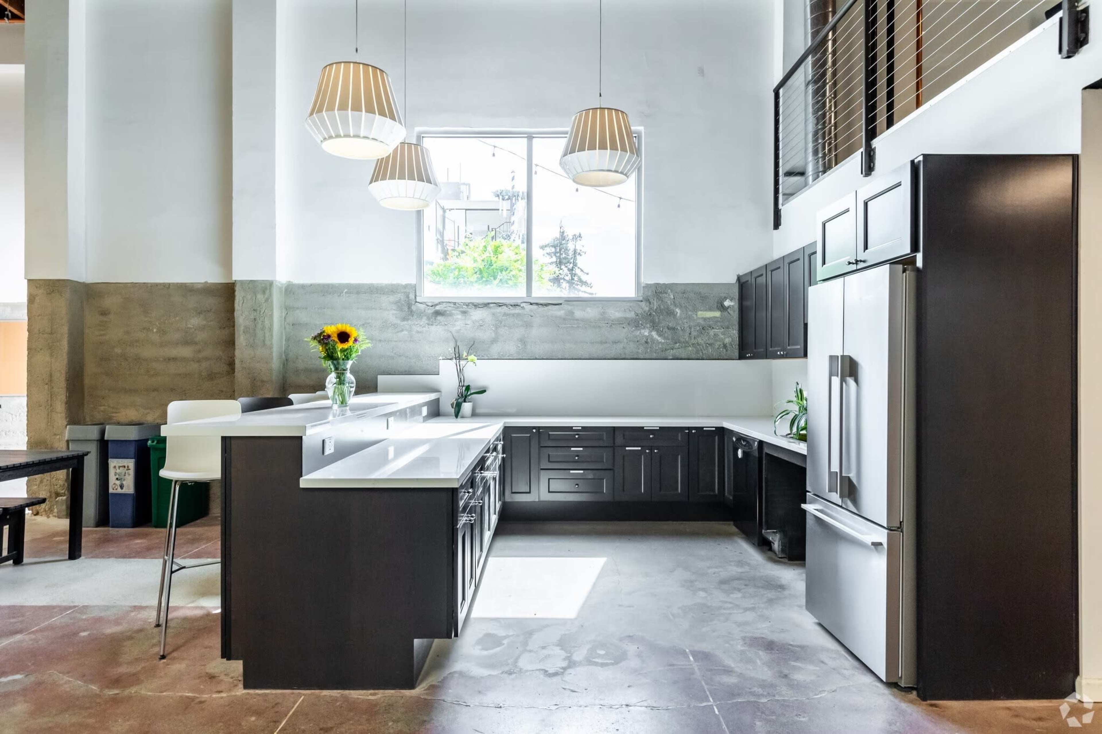The image shows a modern kitchen with dark cabinetry and white countertops, featuring a central island and large windows that allow natural light to enter.