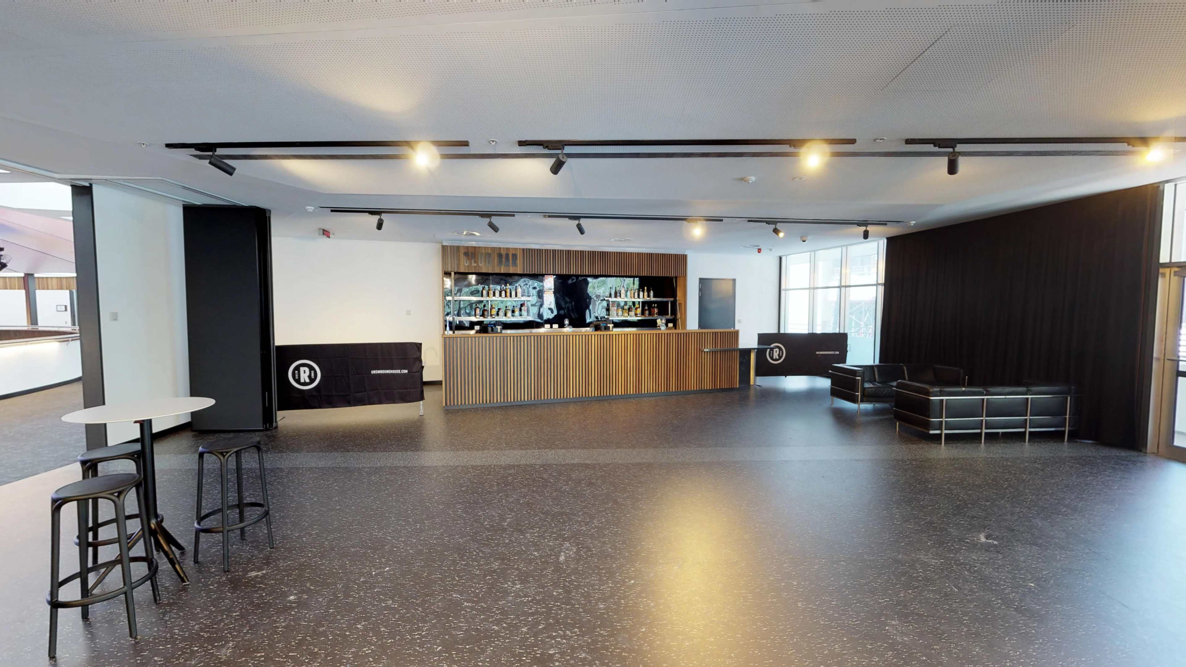 A modern bar area with a wooden counter, shelves behind it filled with bottles, and a seating area featuring a black couch and high chairs.
