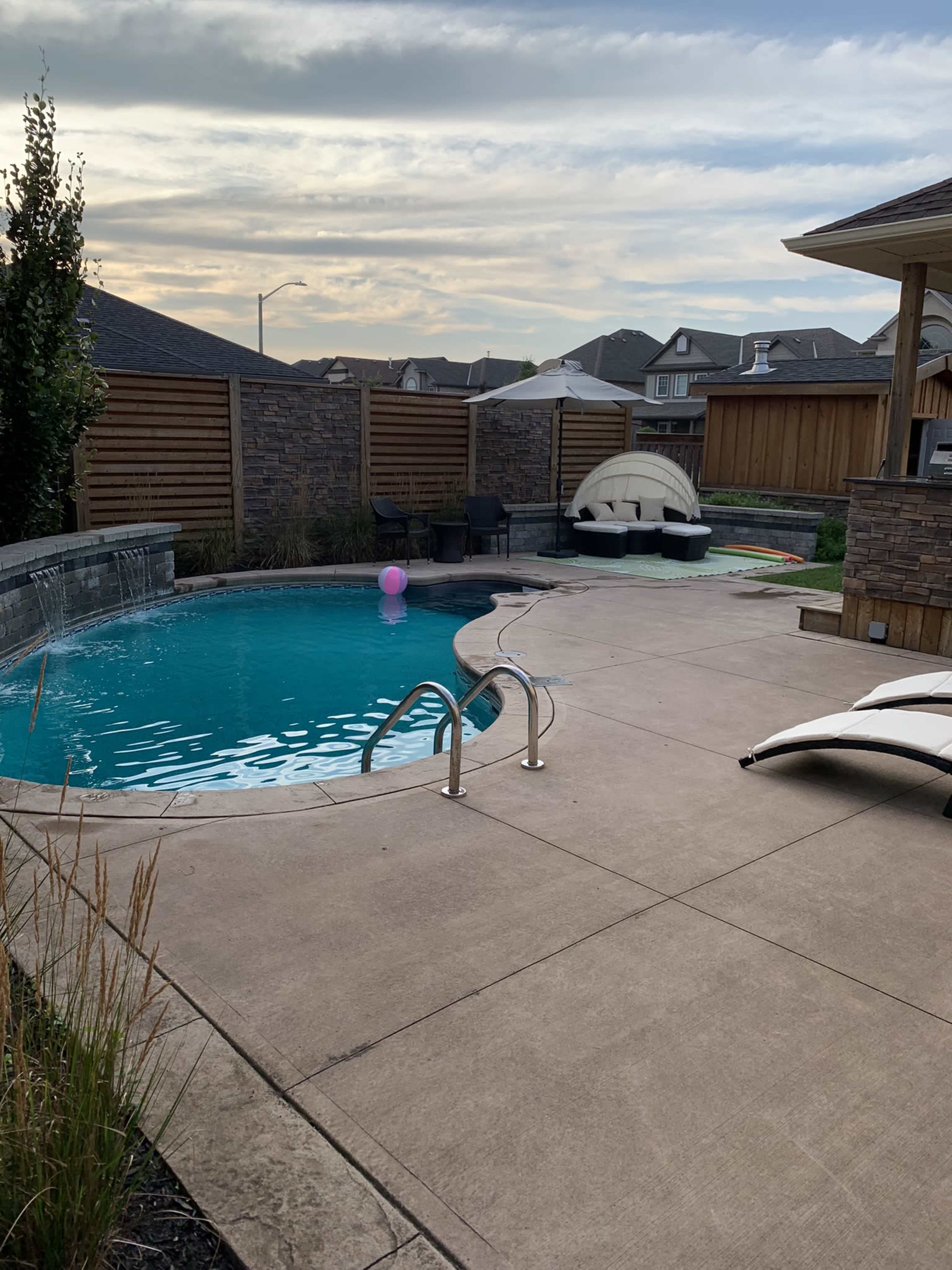 A swimming pool with a slide and lounge chairs is situated in a backyard enclosed by wooden fencing and stone walls.