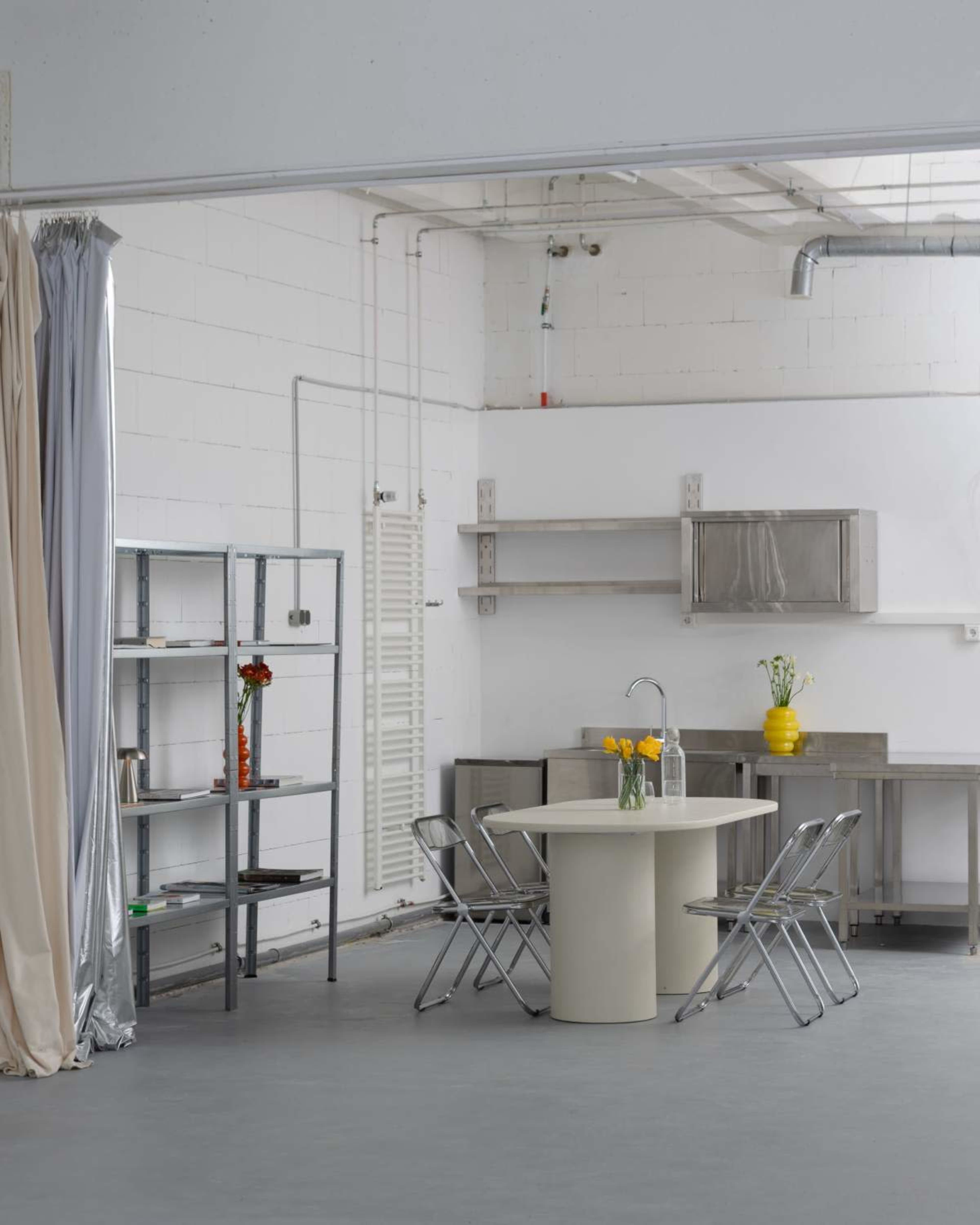 The image shows a spacious kitchen area with a round table and four folding chairs, along with metal shelving and minimal decorative elements.