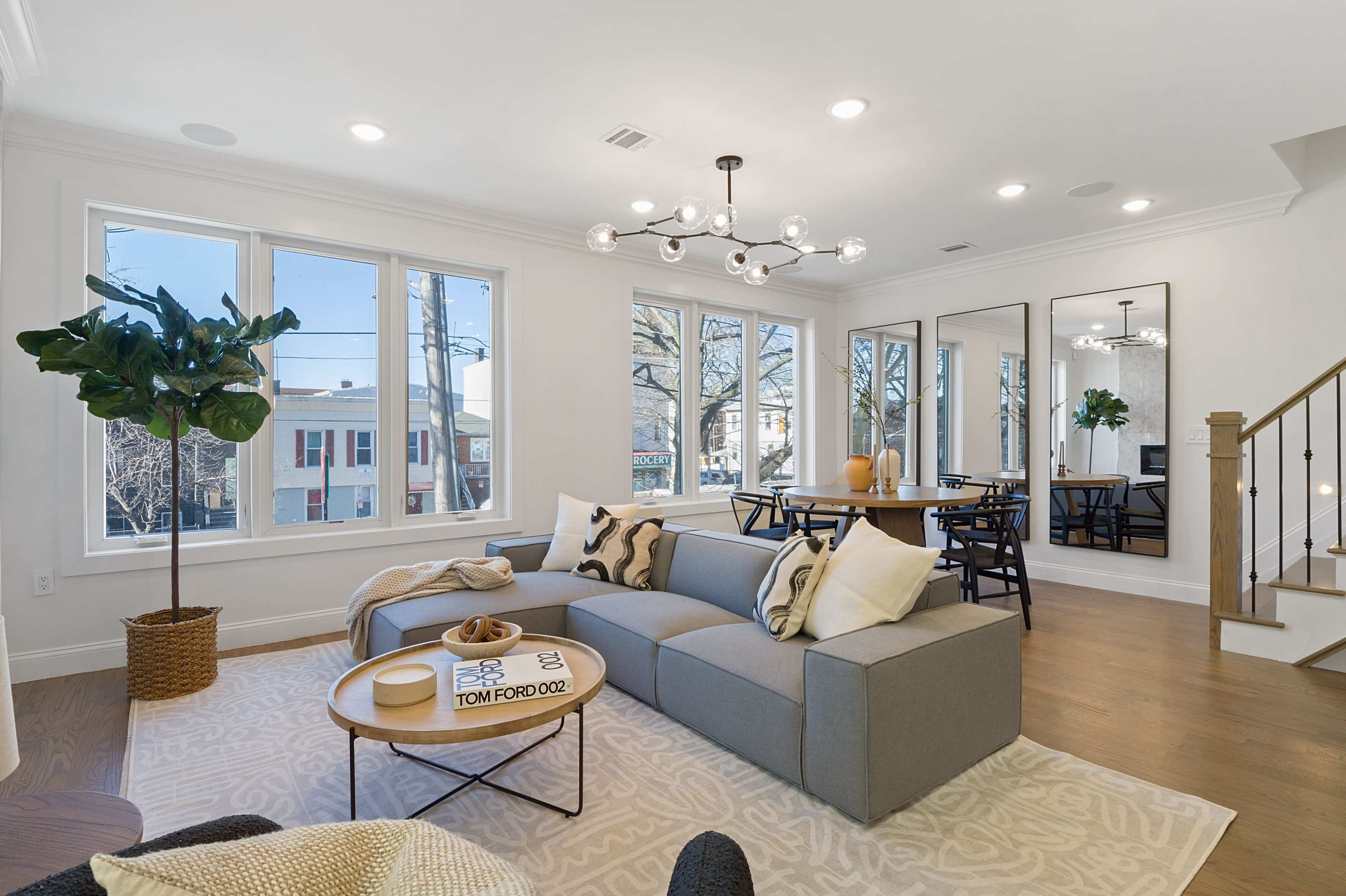 A modern living area featuring a gray sectional sofa, a round coffee table, large windows, and a dining area with a mirror and elegant lighting.