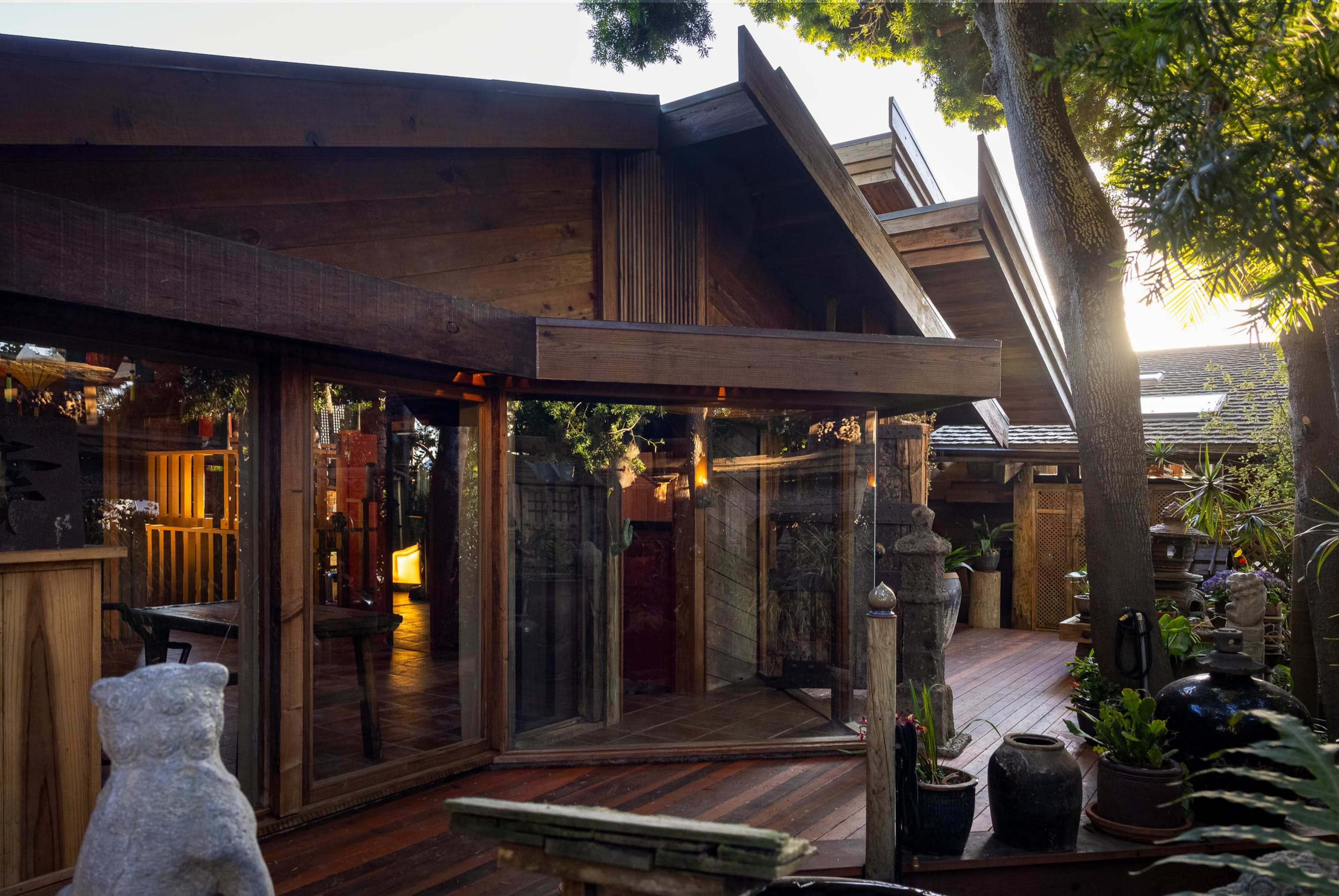 The image shows a modern wooden house with angular roofs and large glass windows, surrounded by greenery and decorated with various potted plants and sculptures.