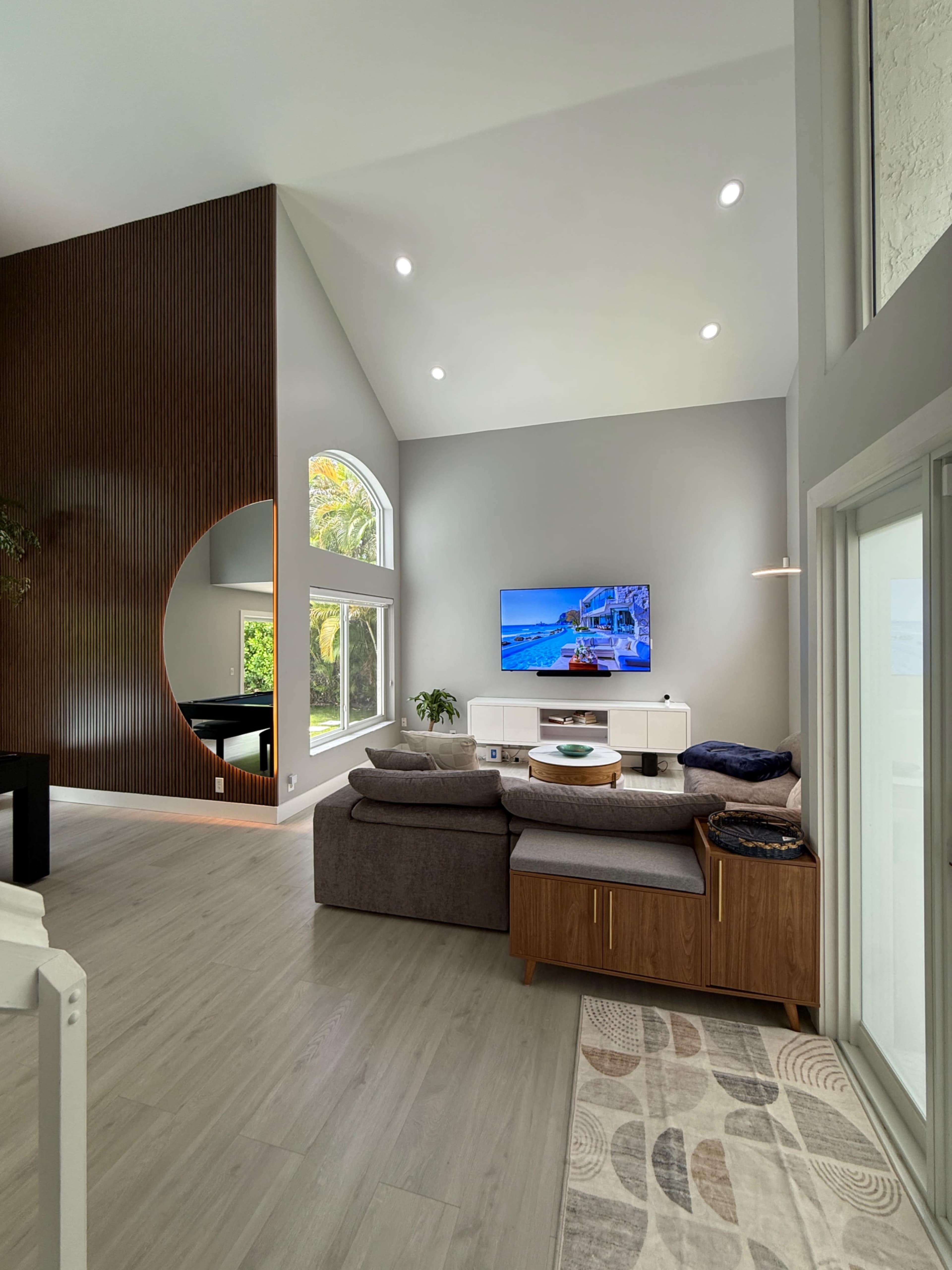 The image shows a modern living room with large windows, a sectional sofa, a wall-mounted TV, and a circular cutout in a feature wall.