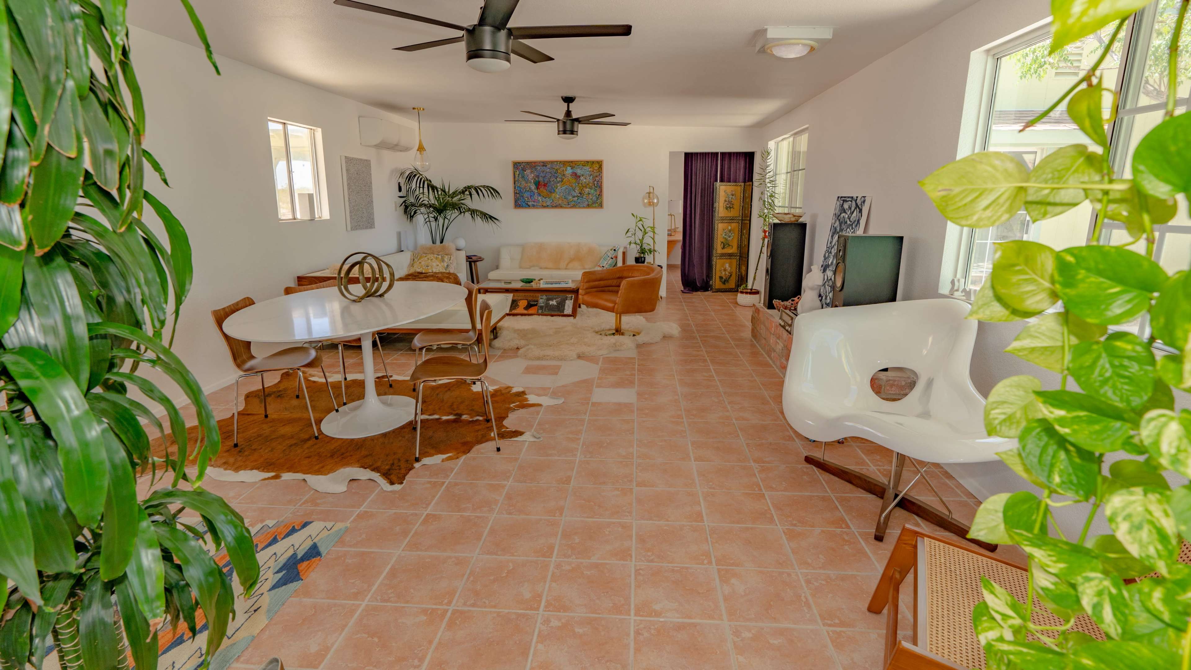 A living room features tiled flooring, a circular dining table, brown seating, and large windows with greenery visible.