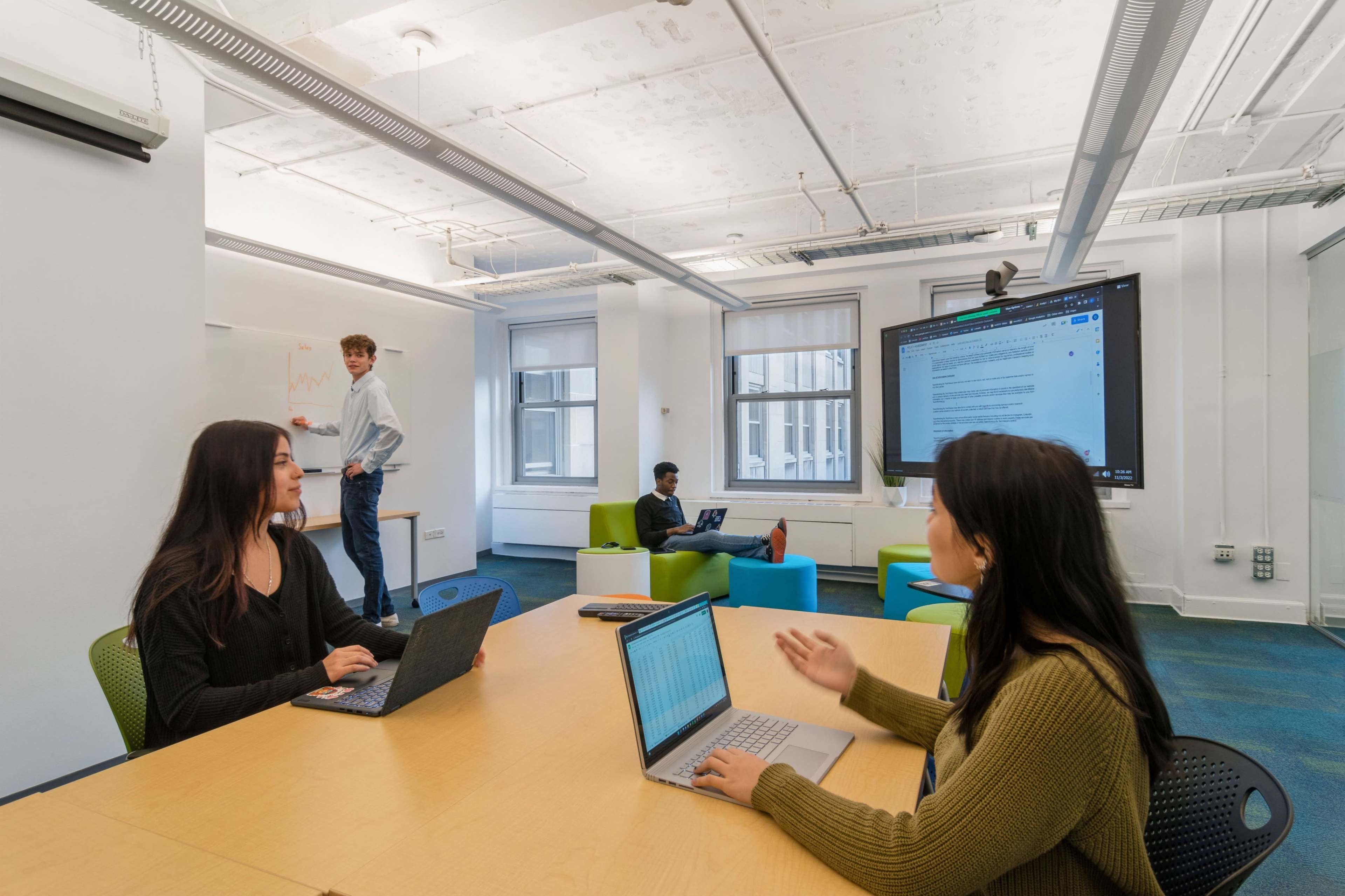 A group of four students engages in discussions and work around a table in a modern classroom, with one student presenting on a whiteboard and others using laptops.