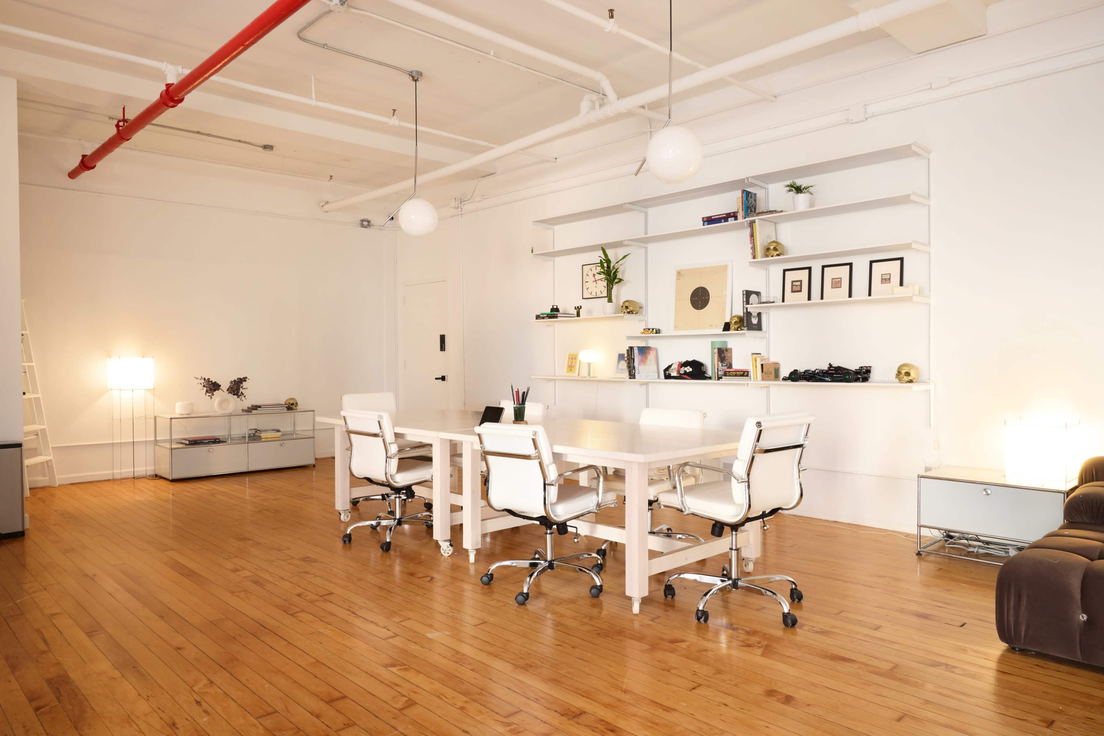 The image shows a modern office space with a long white table surrounded by five white chairs, a bookshelf with decor items, and warm wooden flooring.
