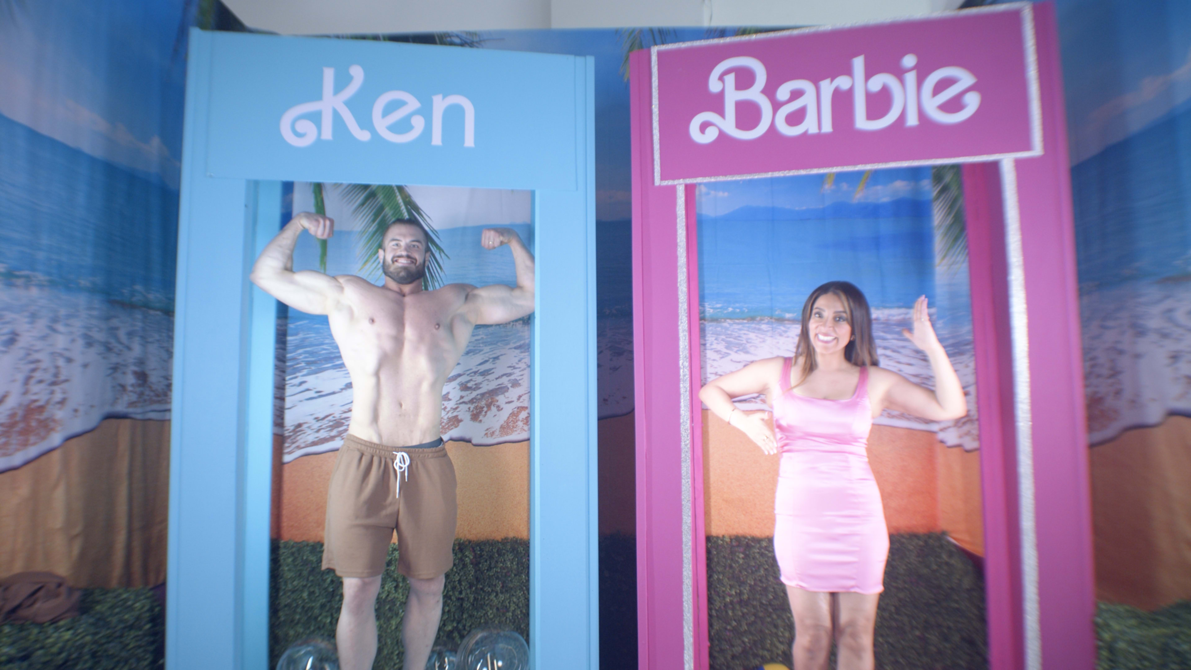 A muscular man poses flexing his arms in a blue booth labeled "Ken," while a woman in a pink dress waves from a pink booth labeled "Barbie."