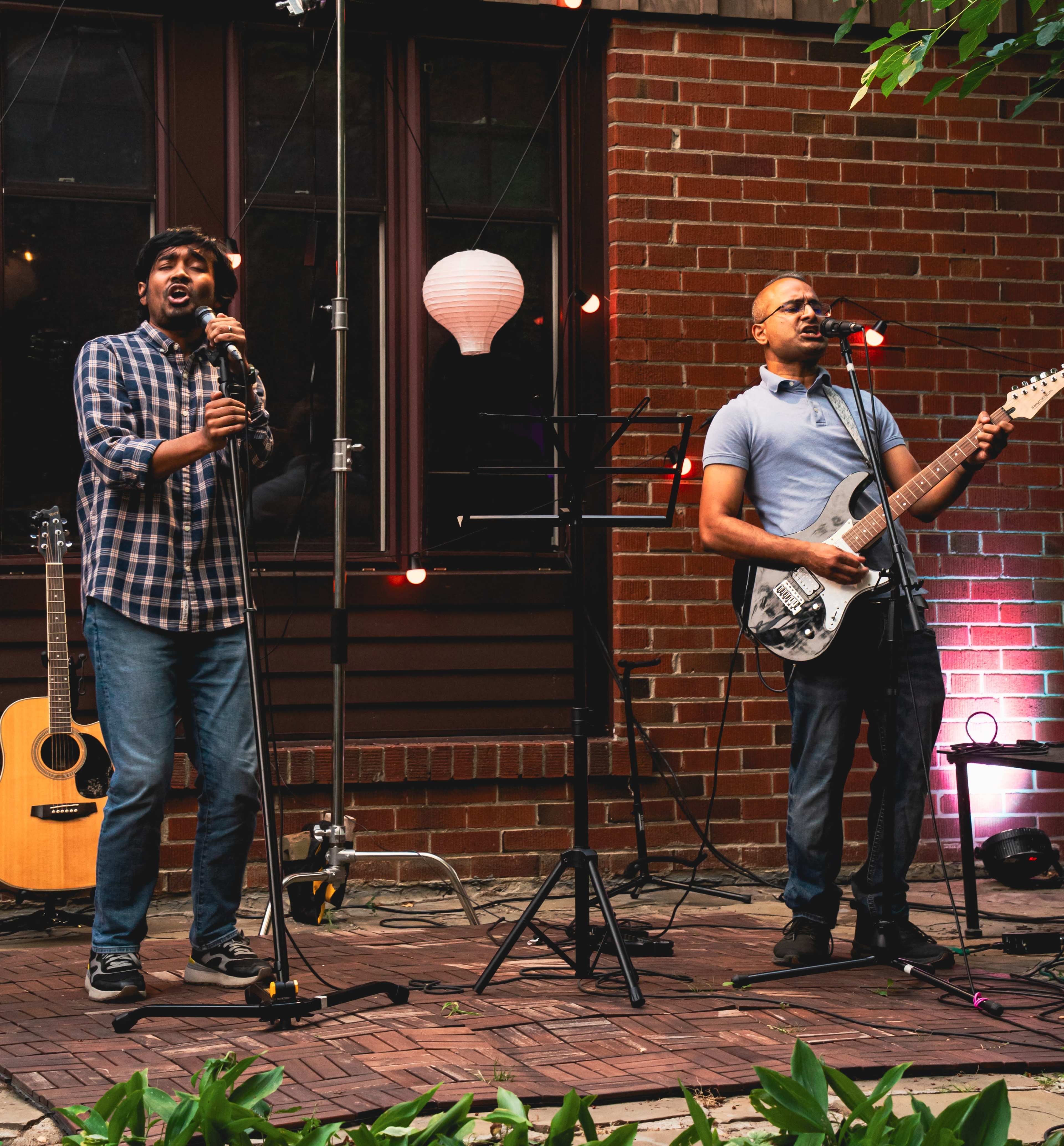 Two male musicians perform on a small outdoor stage with a guitar and a microphone, surrounded by red brick walls and decorative lights.