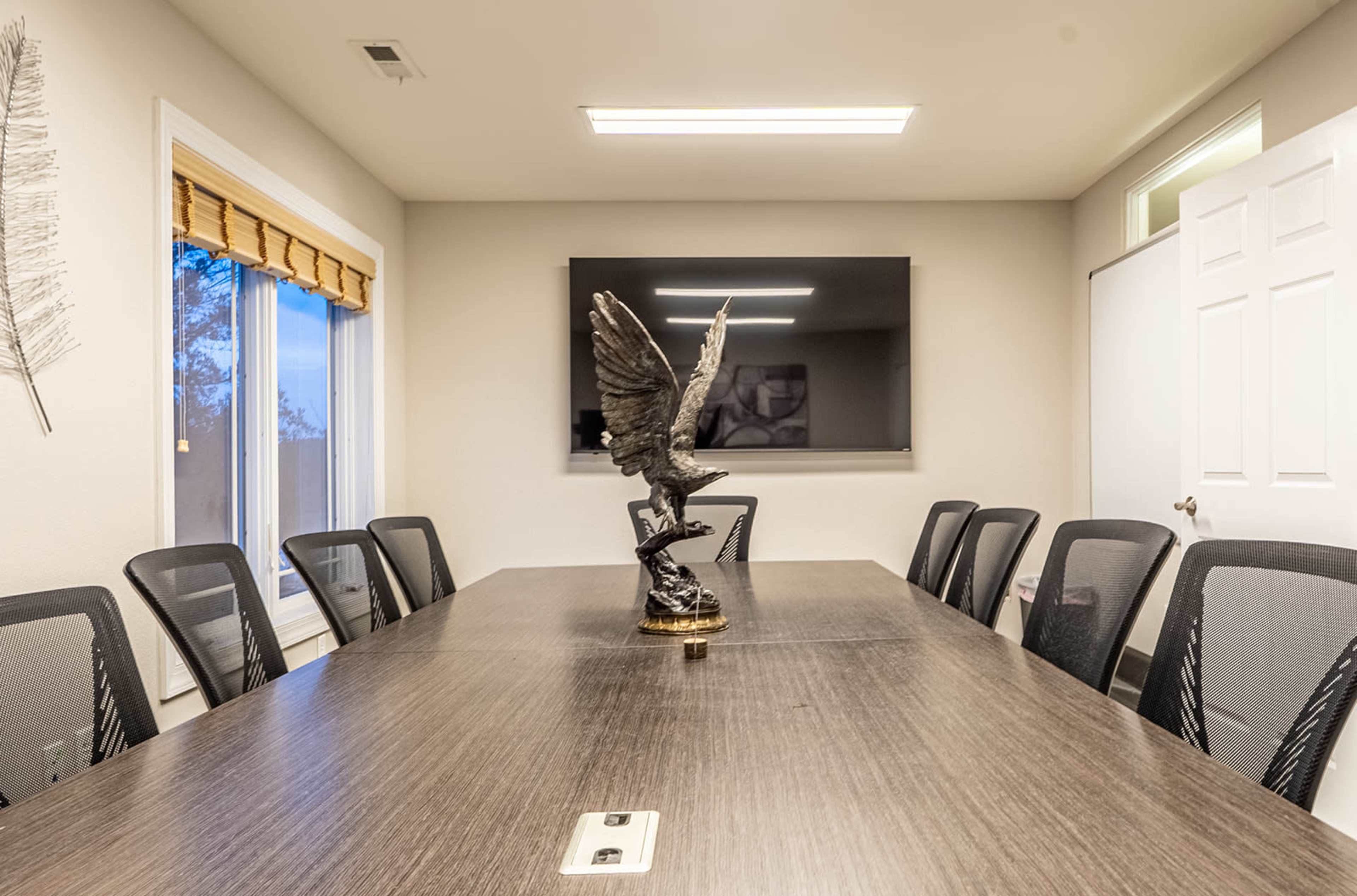 A conference room features a wooden table surrounded by black mesh chairs, with a decorative eagle statue at the center and a television mounted on the wall.