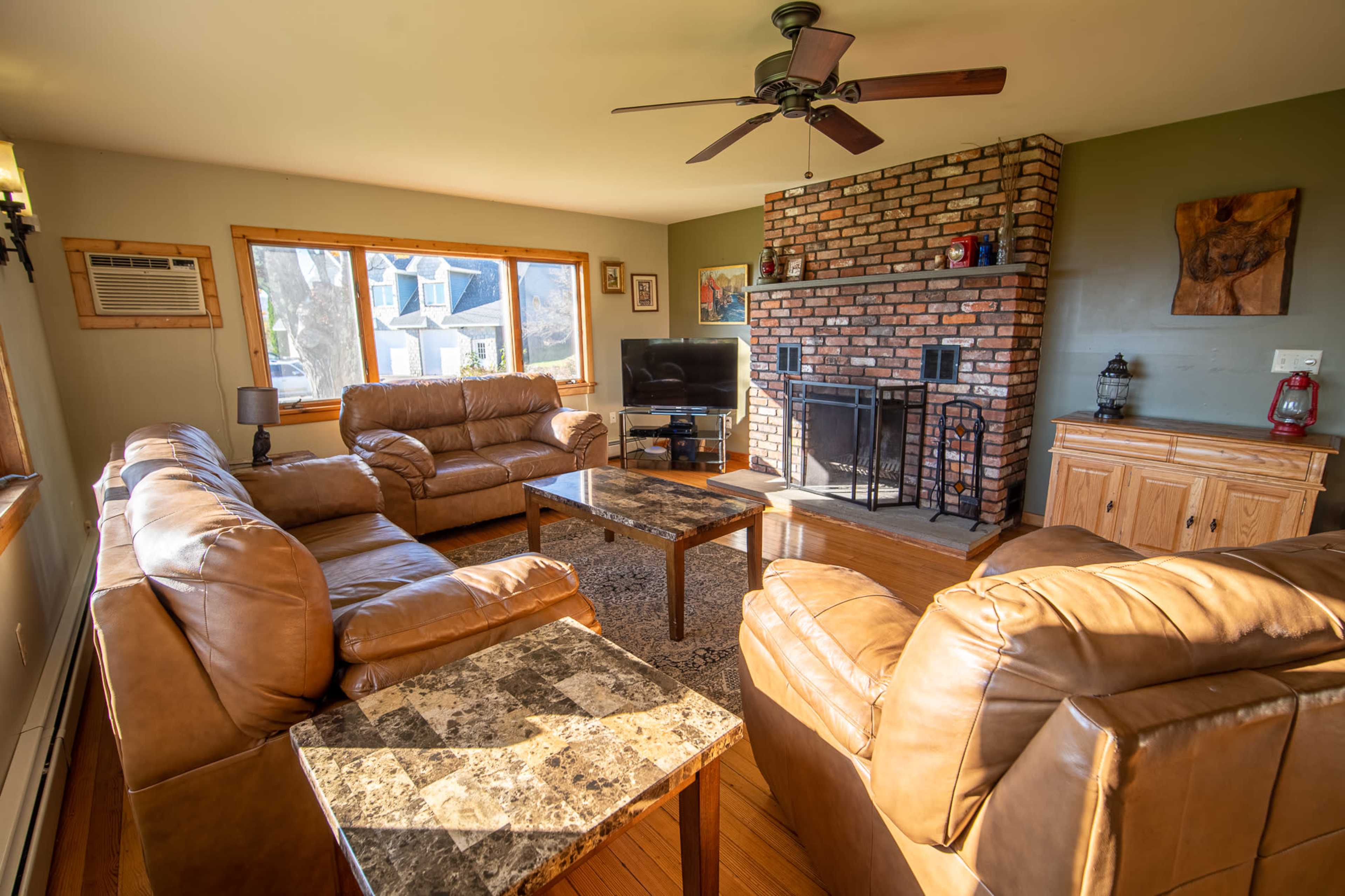 A living room furnished with leather sofas, a coffee table, and a brick fireplace, with a large window providing natural light.