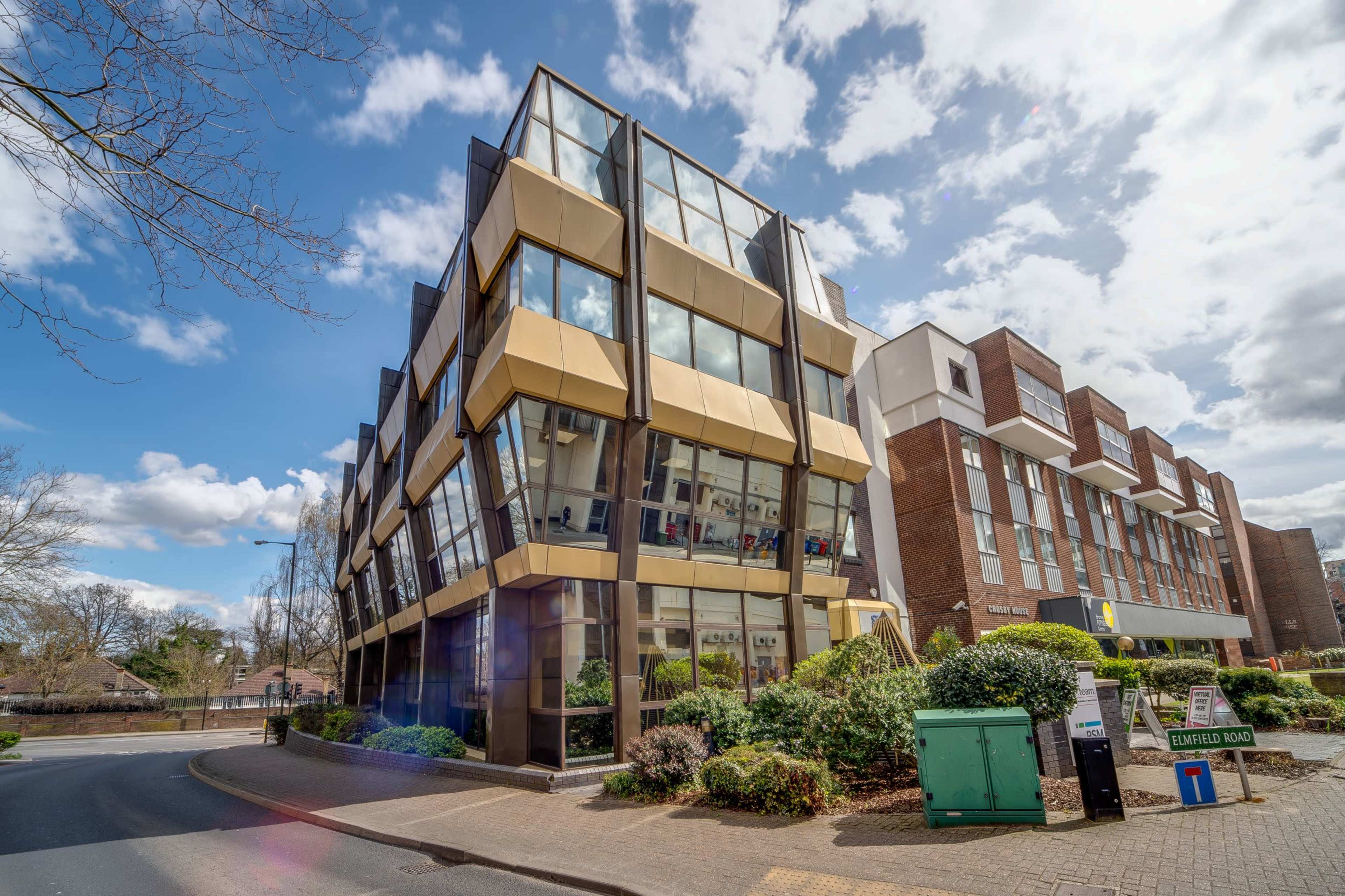A modern, multi-story building with large glass windows and a gold-colored facade is situated beside a landscaped area and a brick structure.
