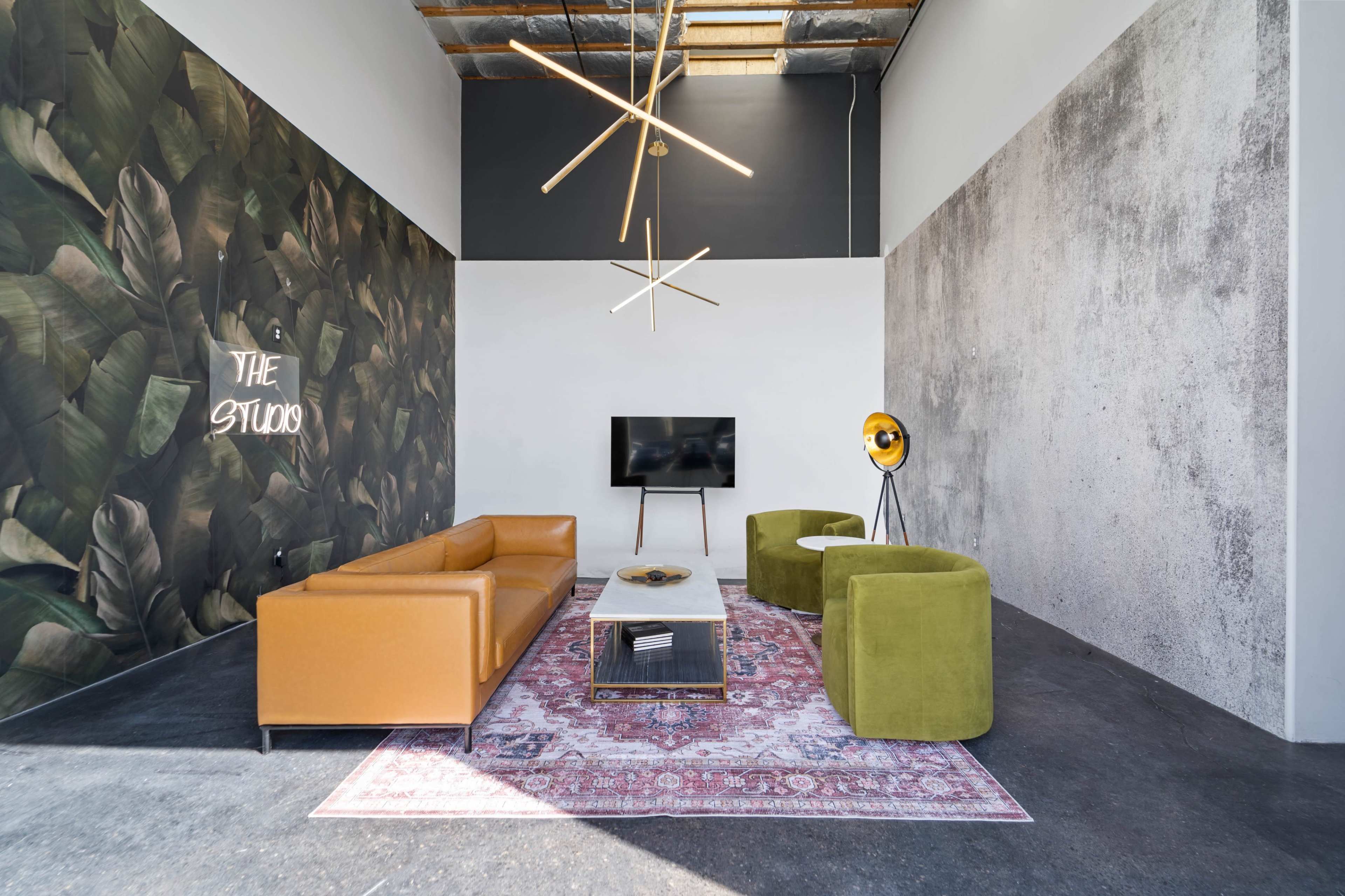 The image showcases a modern studio space with a brown leather sofa, green accent chairs, a patterned rug, and a television mounted on the wall, set against a backdrop of botanical and concrete textures.
