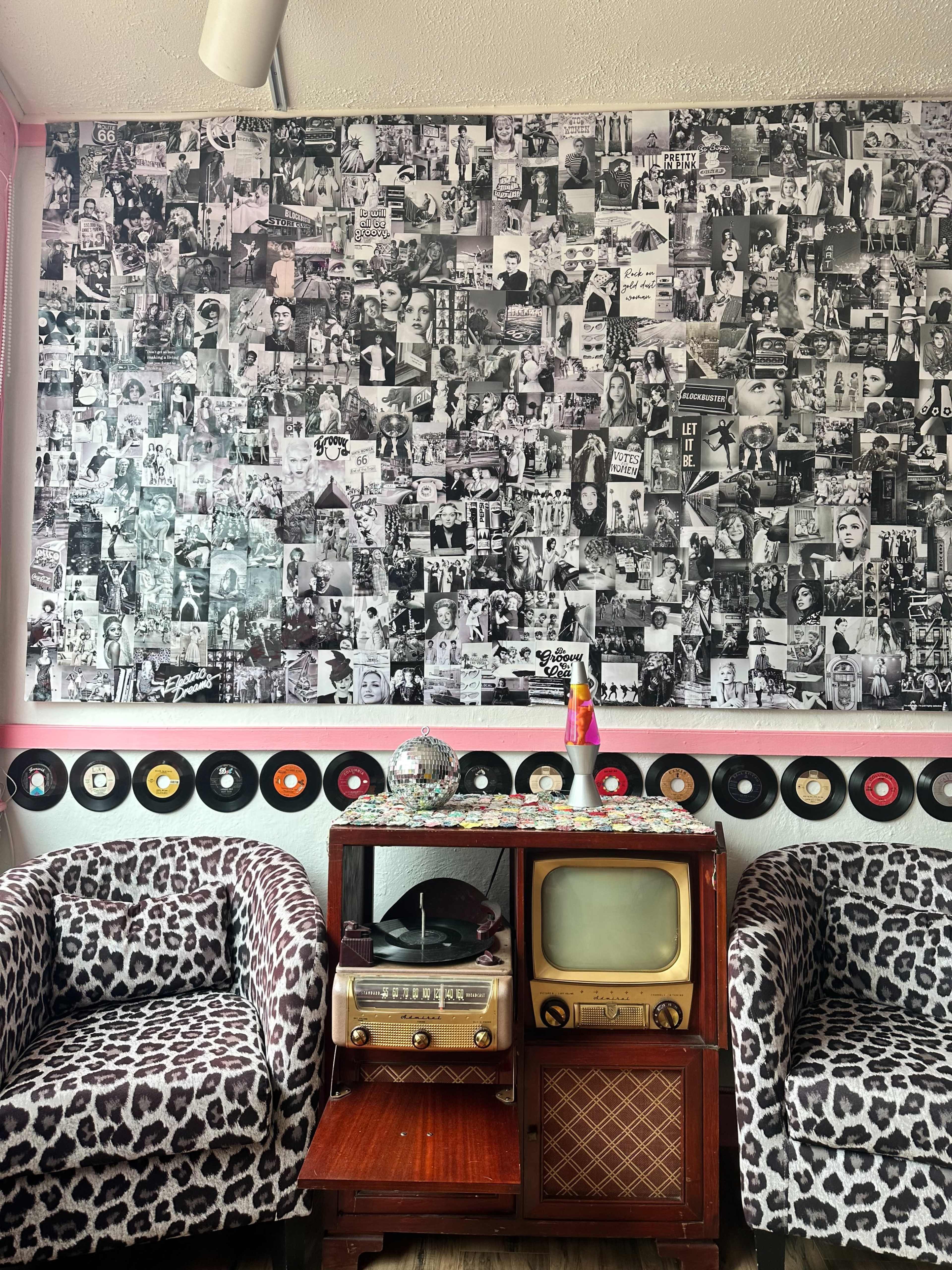 A room with two leopard-print chairs flanking a vintage wooden cabinet that holds an old television and a record player, set against a wall covered with a collage of black-and-white photographs.