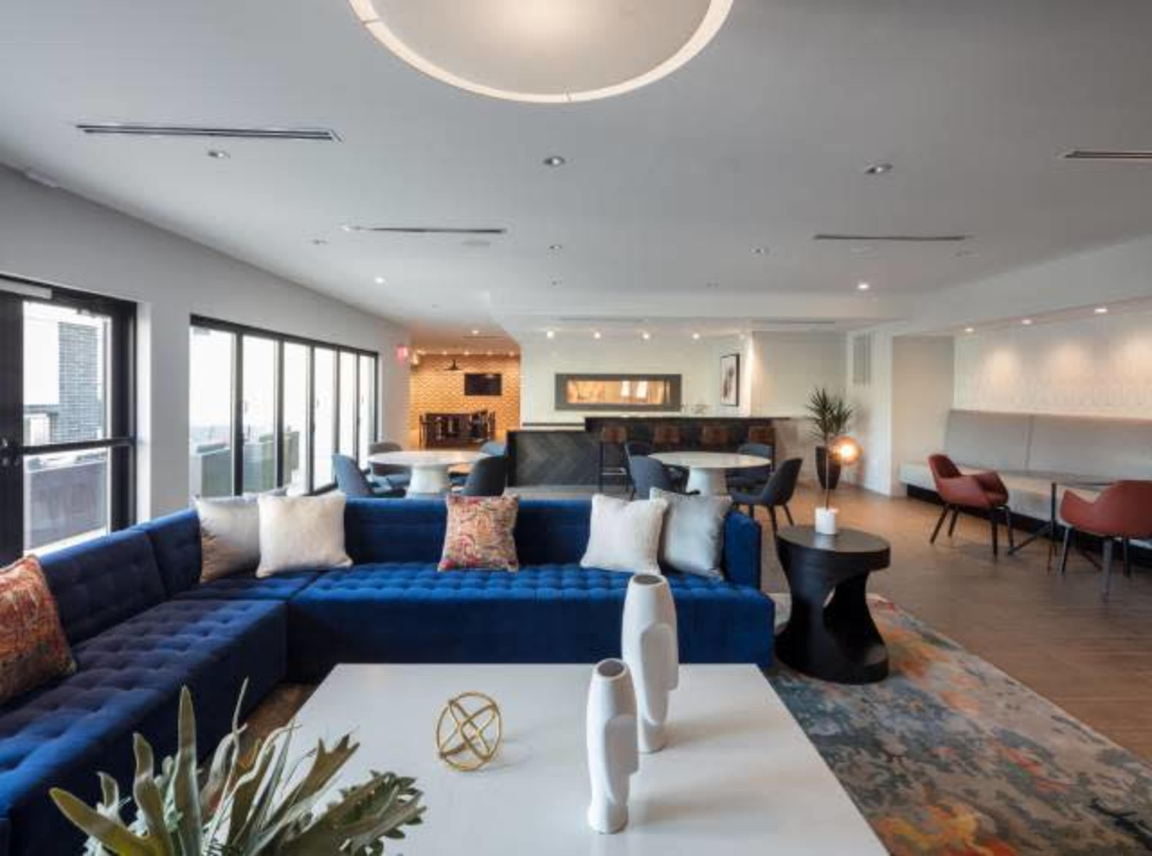 A modern lounge area features a large blue sectional sofa, a minimalist white table, and various seating arrangements with large windows.