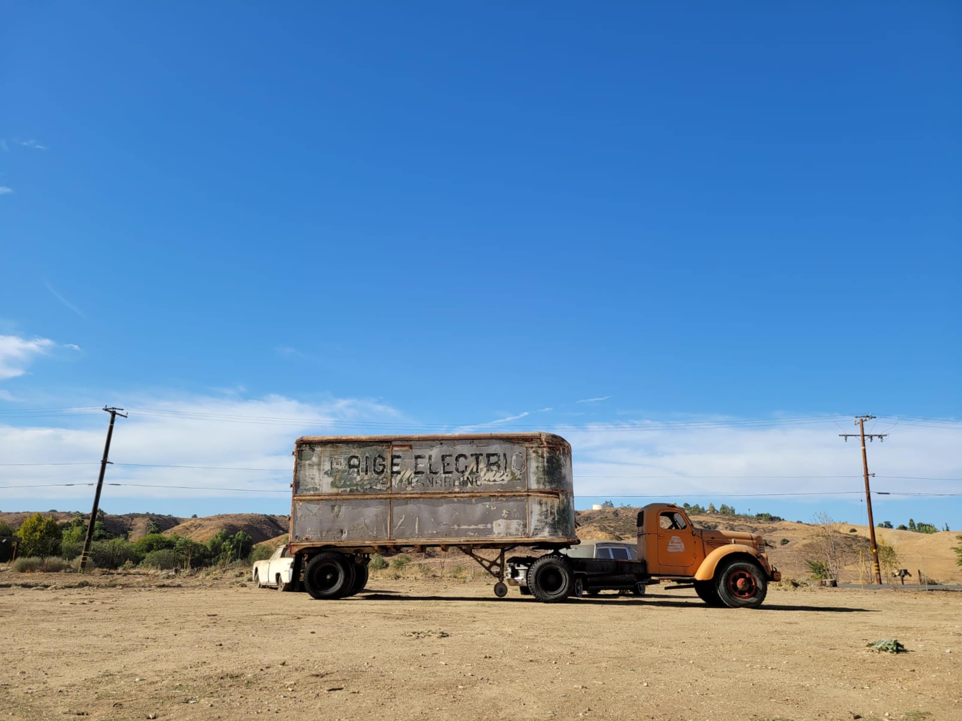 An old, rusty truck with a large trailer parked on a dirt lot under a blue sky with distant hills.