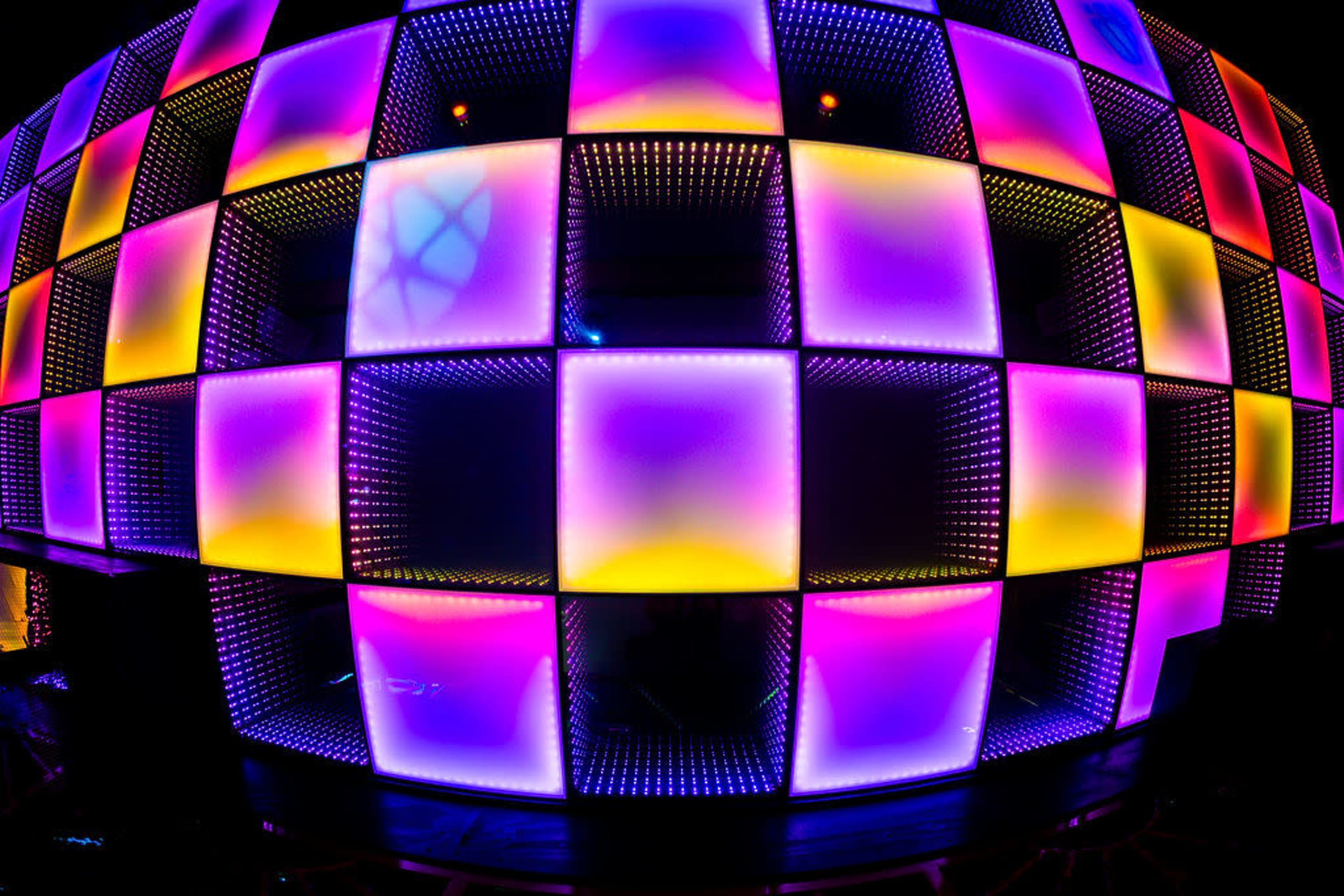 The image displays a large, illuminated wall composed of square panels that alternate in vibrant colors of purple, pink, and yellow.