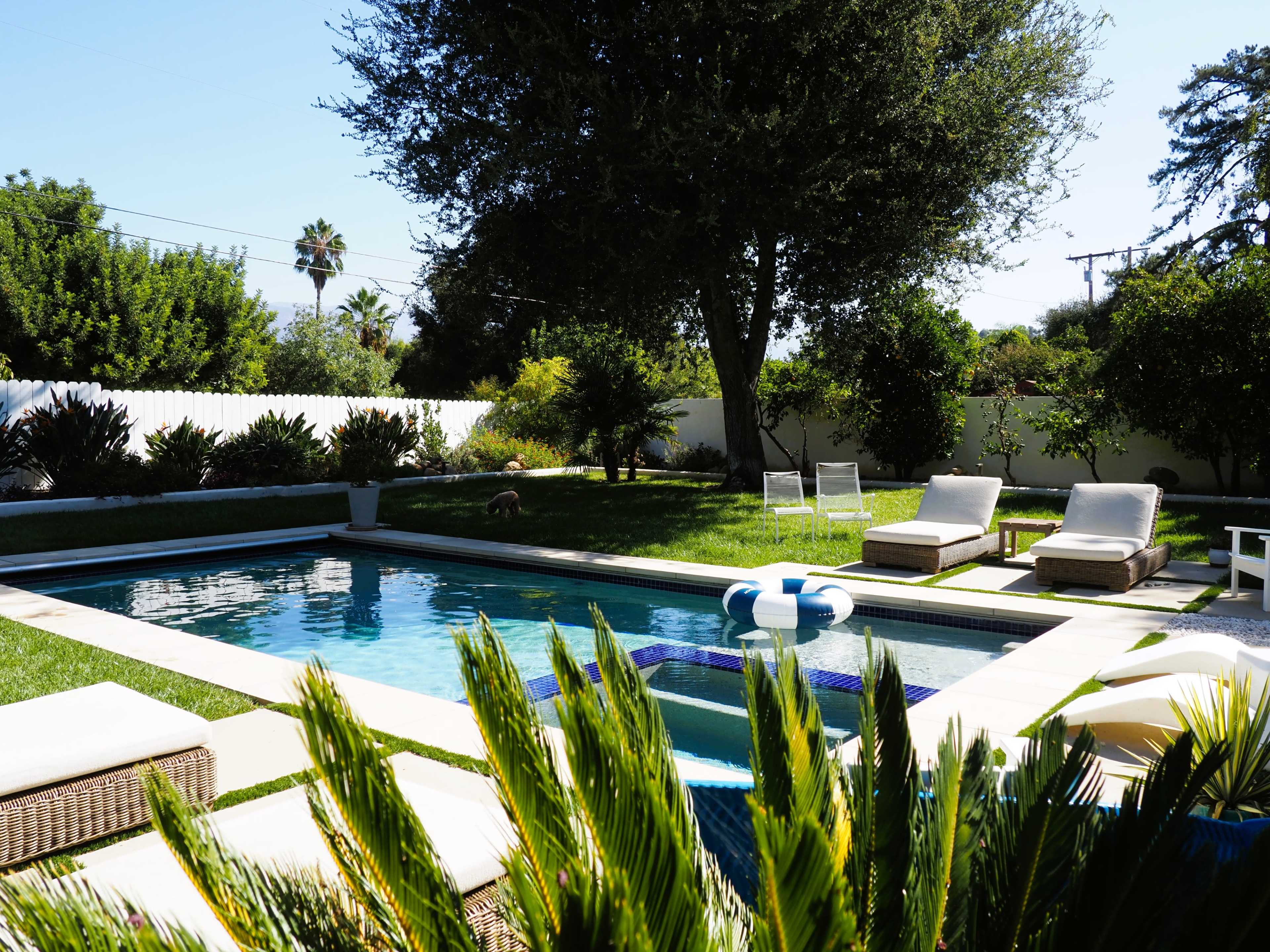 The image shows a swimming pool surrounded by greenery, lounge chairs, and a patio in a residential backyard.