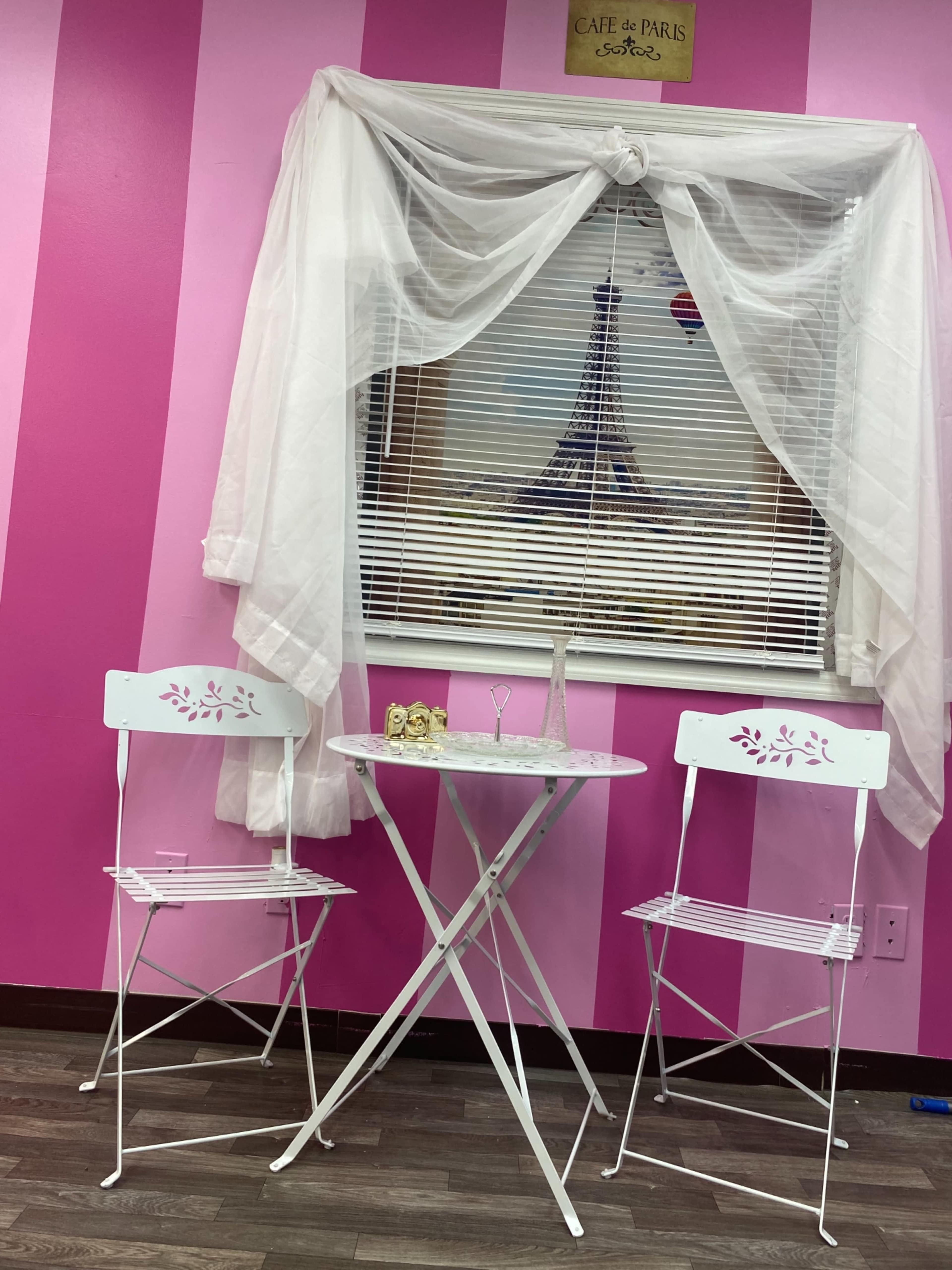 A small, round table with two white chairs is set up in front of a window draped with white fabric, against a background of pink striped walls.