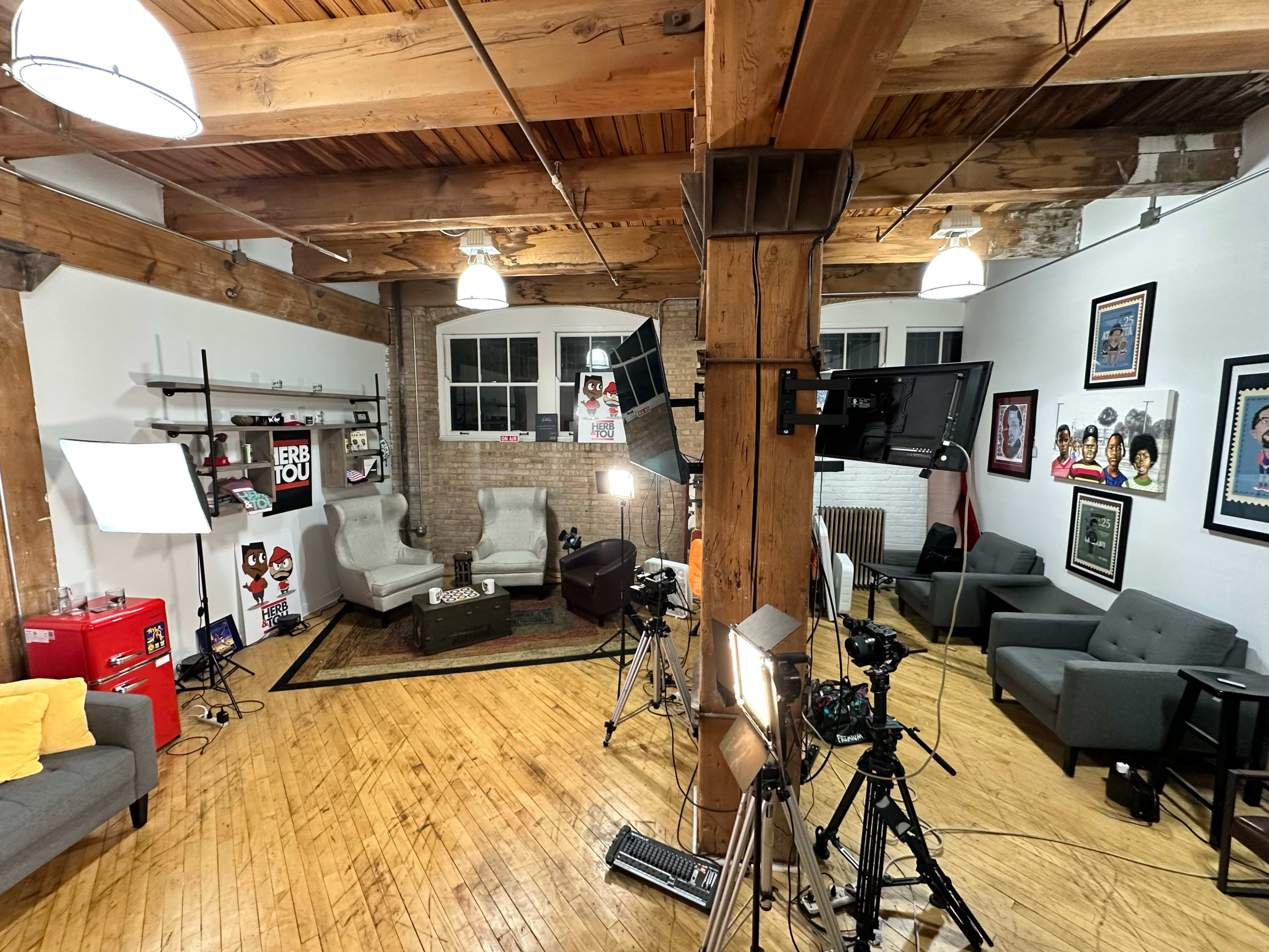 The image shows a well-lit studio setup with cameras, lighting equipment, and a cozy seating area featuring two armchairs, a couch, and various decorative items on the walls.