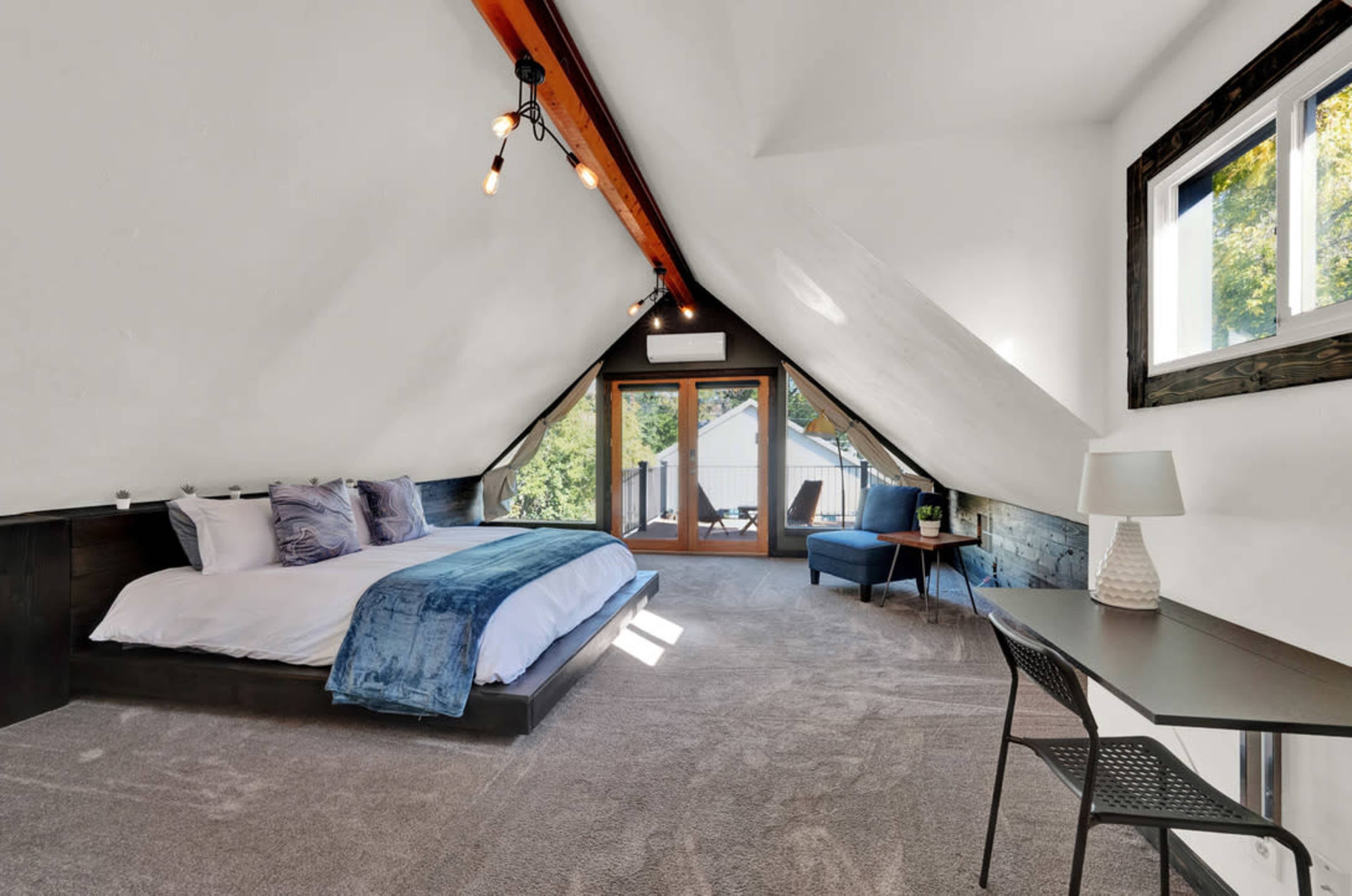 A spacious attic bedroom with a large bed, a desk, and a seating area, featuring sloped ceilings and large windows.