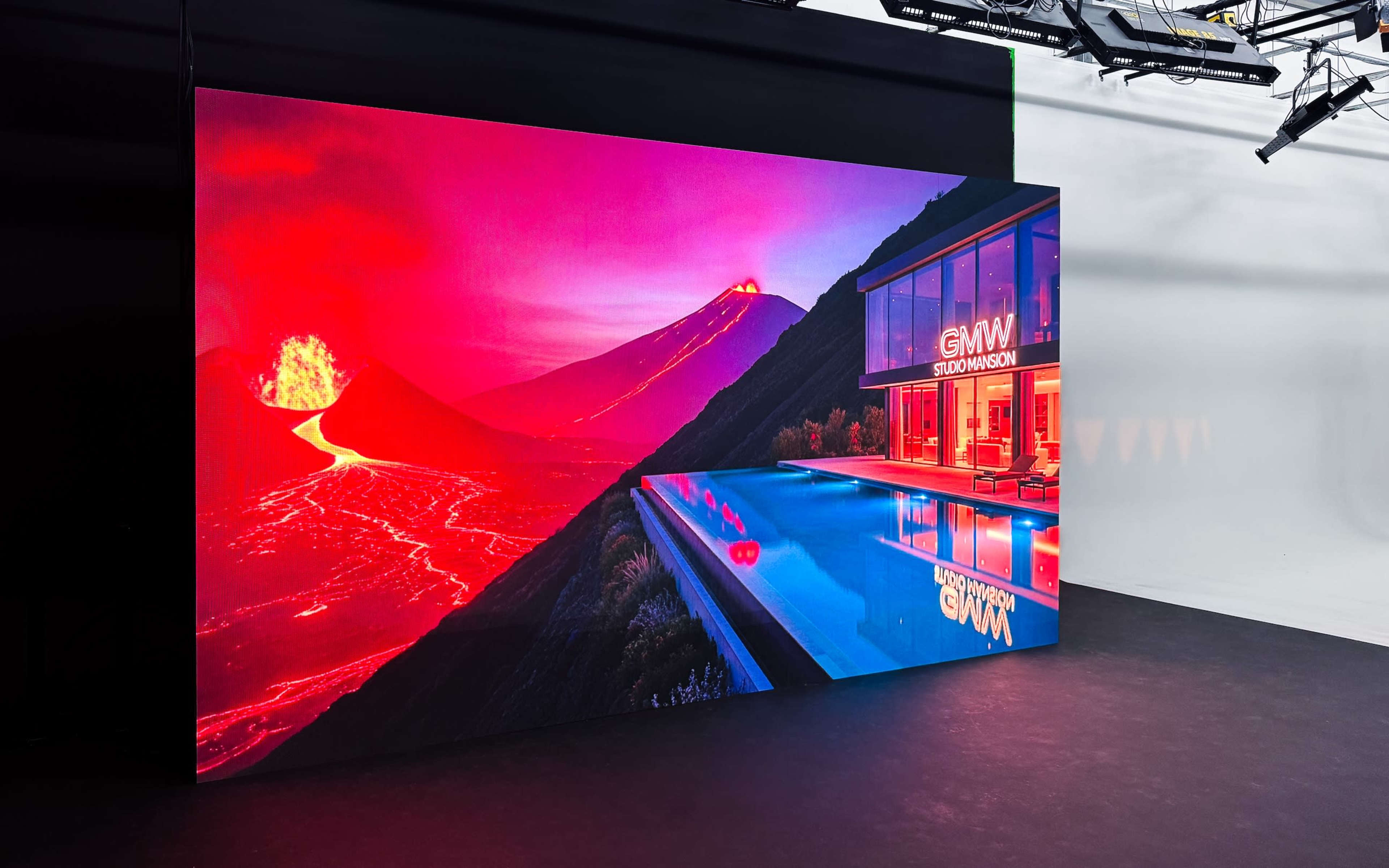 The image features a large, vivid display showing a volcanic landscape with lava flowing, alongside a modern building with a pool reflecting the scene.