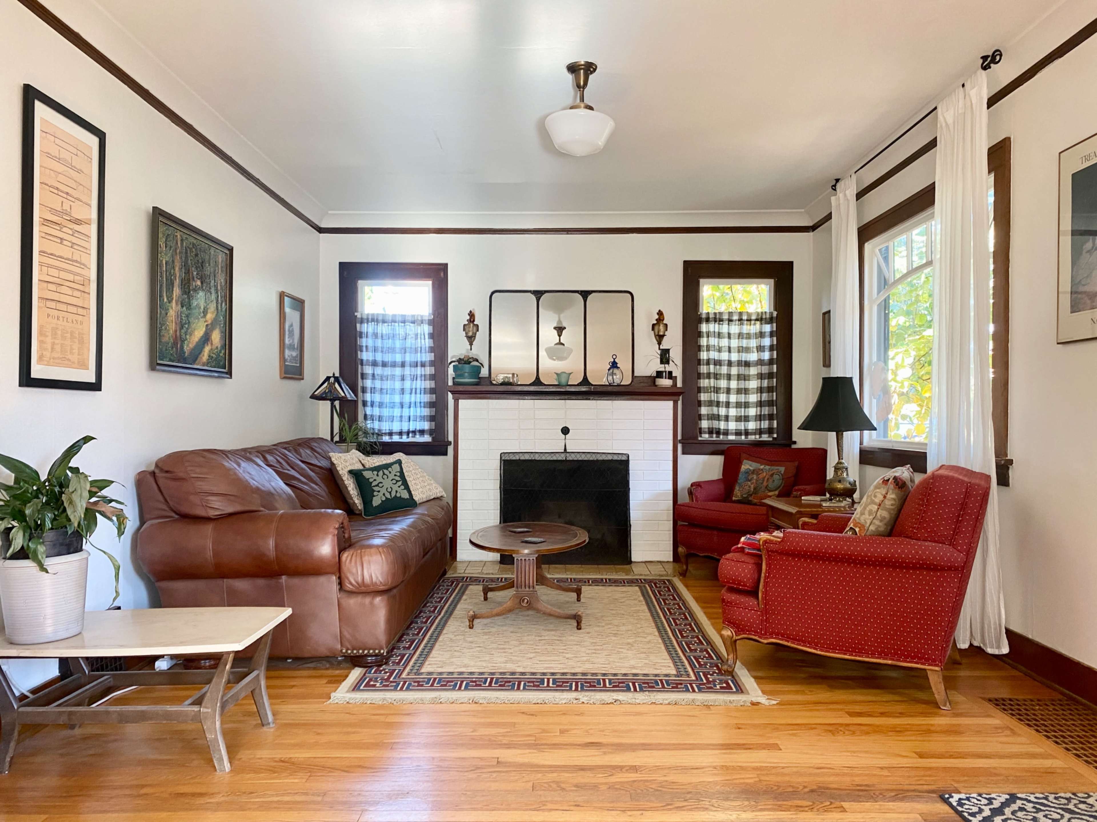 A living room features a brown leather sofa, a red armchair, a coffee table, and a fireplace with decorative items on the mantel, all surrounded by hardwood flooring and natural light from the windows.