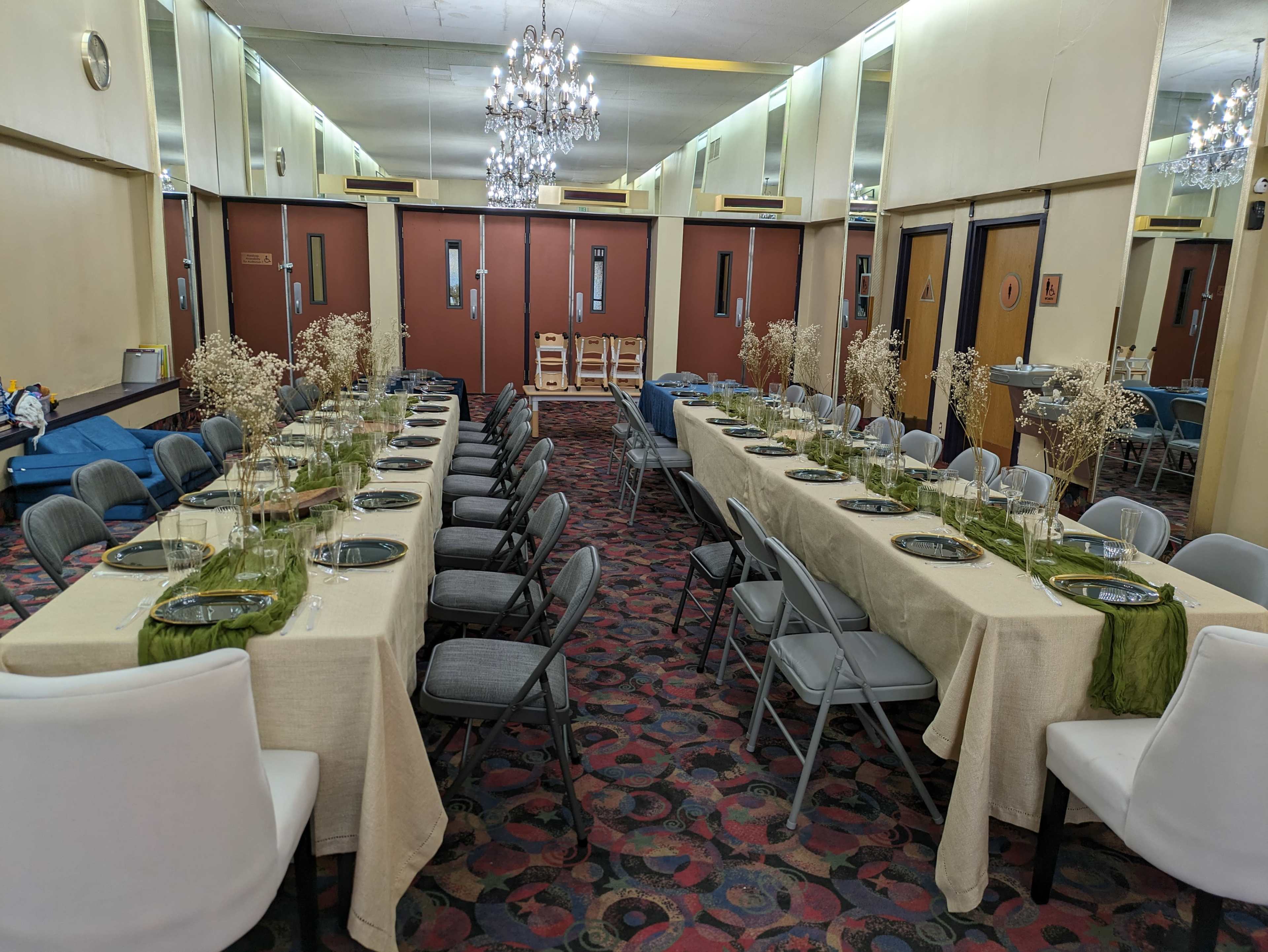 A symmetrical arrangement of two long tables is set for a formal gathering, adorned with green and clear glass plates and decorated with dried flowers, in a room with mirrors and chandeliers.