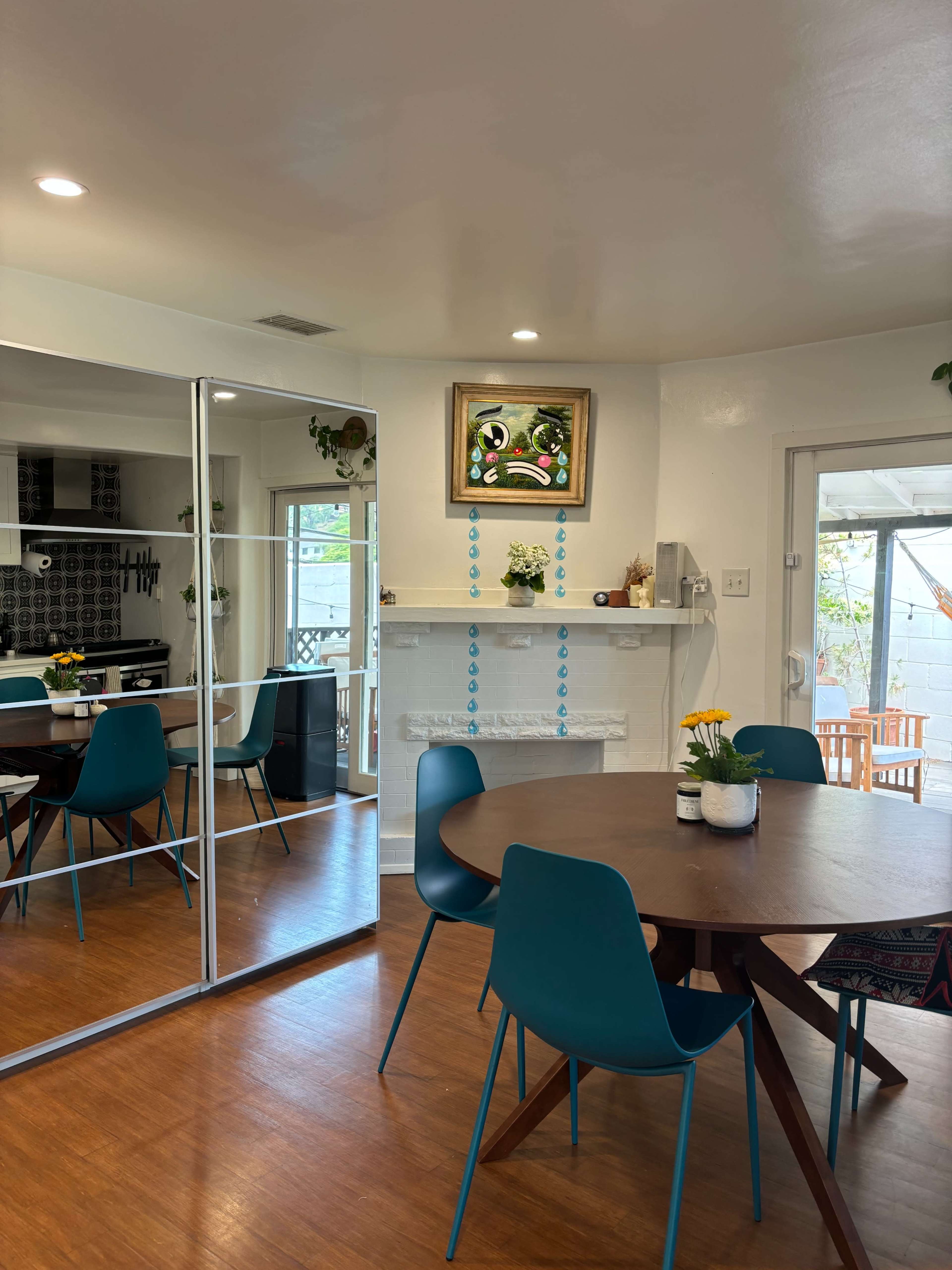 The image shows a modern dining area with a round wooden table surrounded by blue chairs, a mirrored wall, and a decorative fireplace with artwork above it.