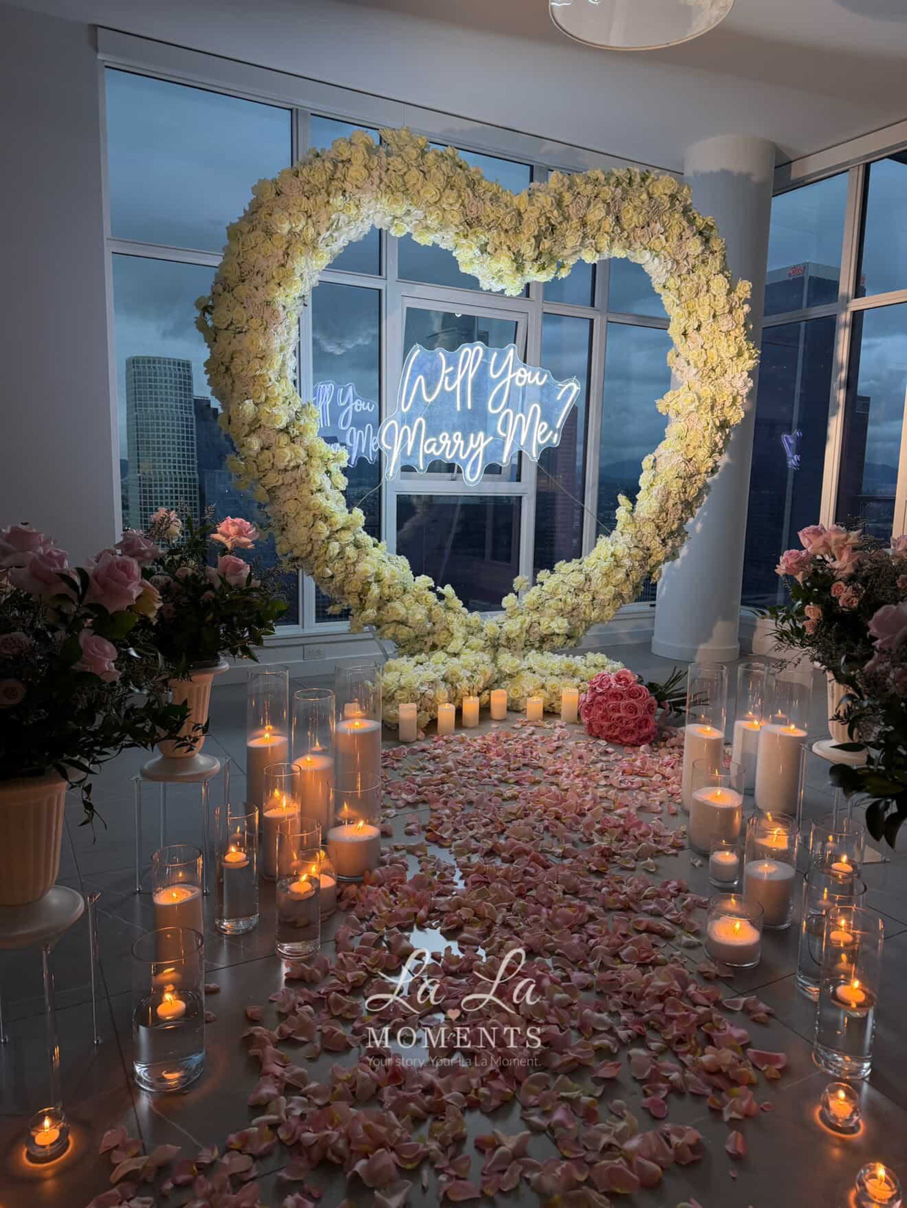The image depicts a romantic proposal setup featuring a heart-shaped arrangement of flowers, a neon sign that reads "Will You Marry Me?" and a path lined with candlelit glass holders and rose petals leading to the display.