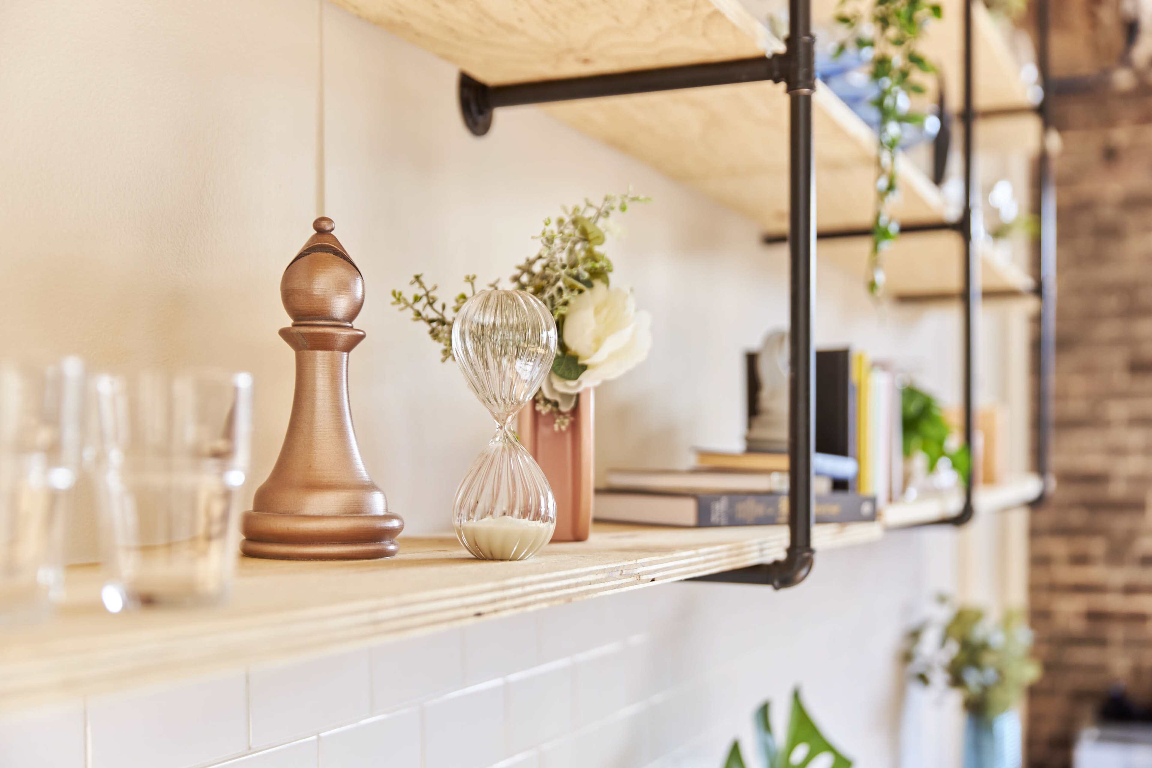 A wooden shelf displays a wooden piece, an hourglass, a flower vase, books, and glasses against a textured wall.