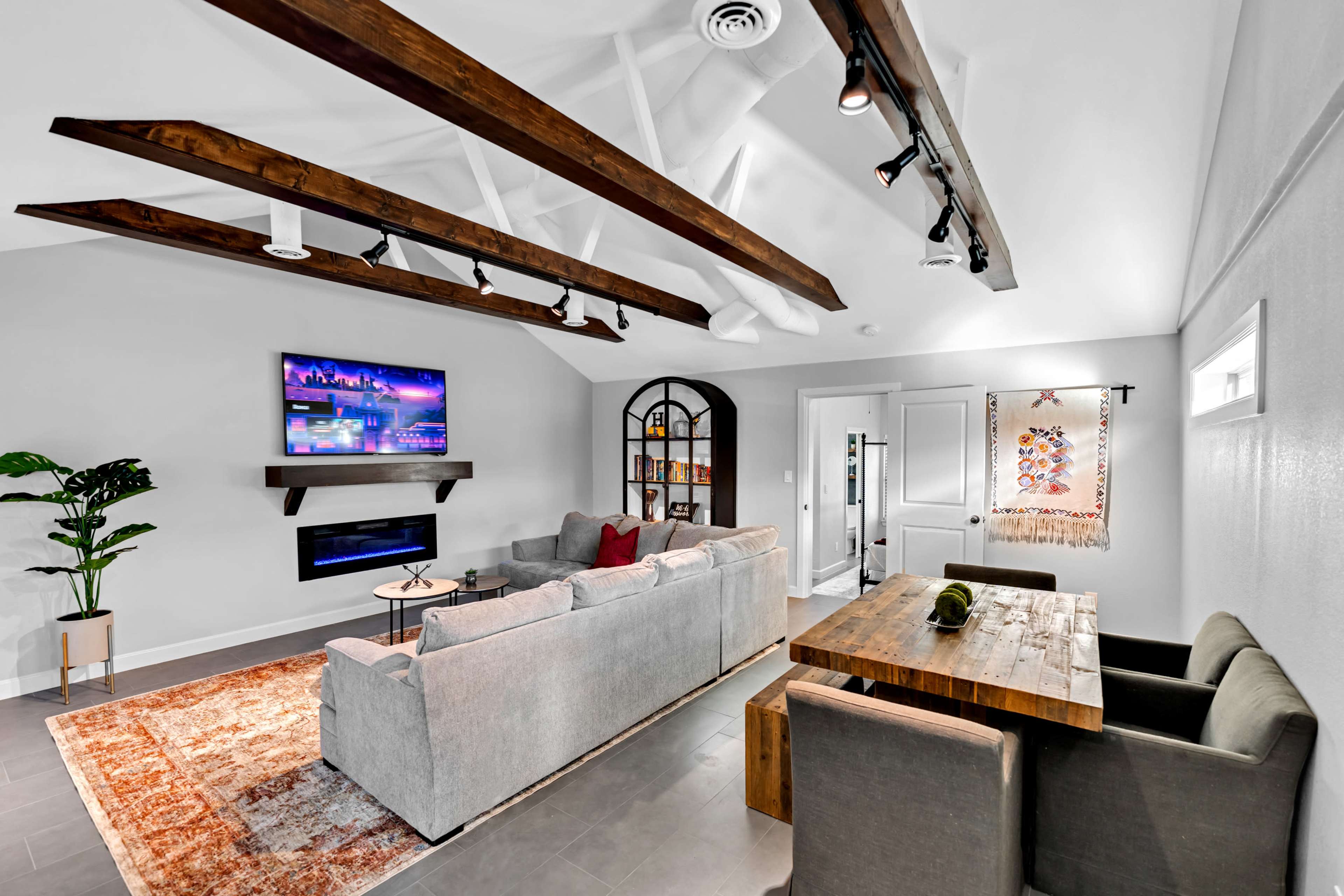 A modern living room featuring a gray sectional sofa, a wooden dining table, a wall-mounted television, and beams across the ceiling.