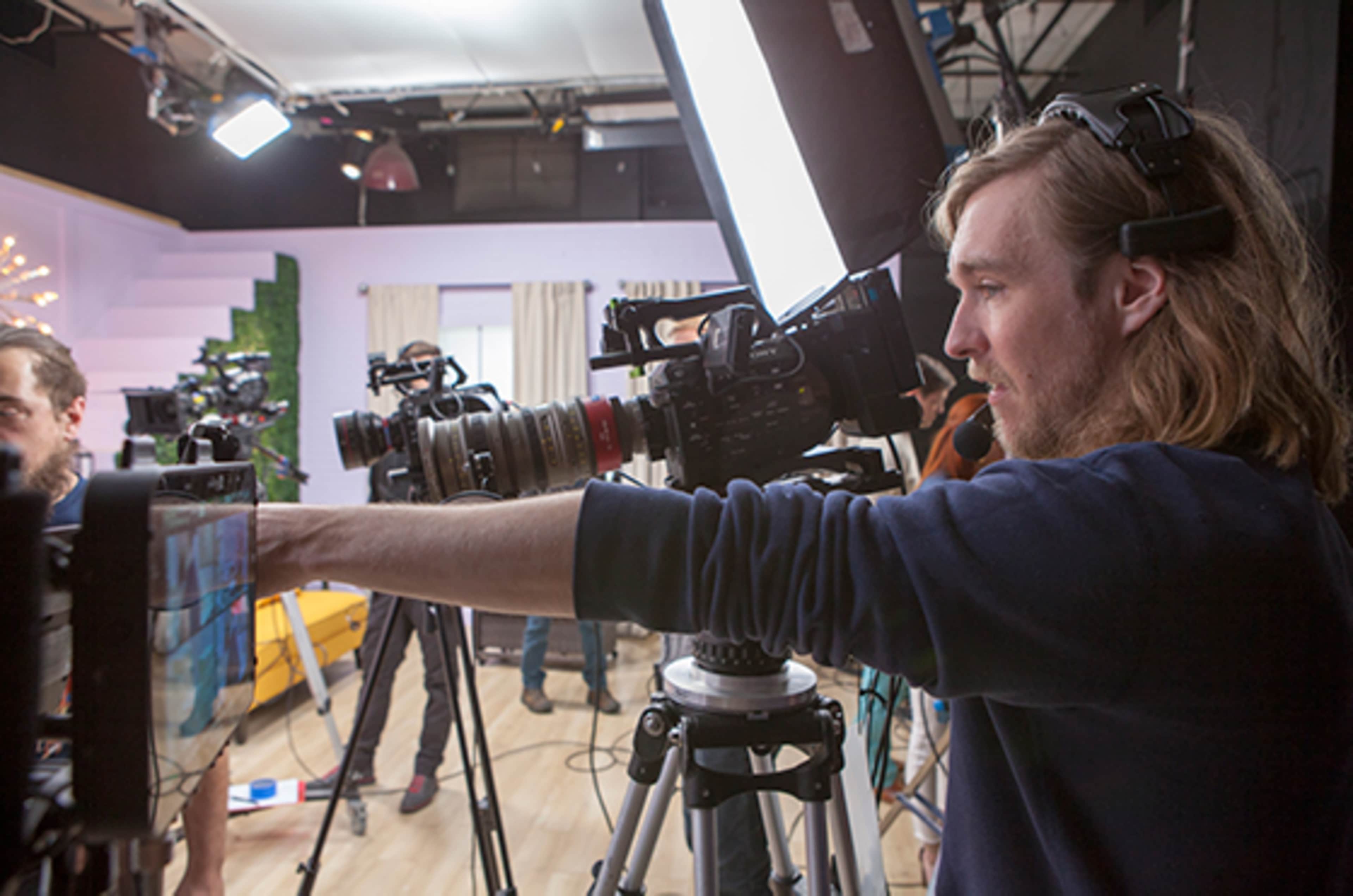 A crew member operates a camera on set, surrounded by other filming equipment and crew members.