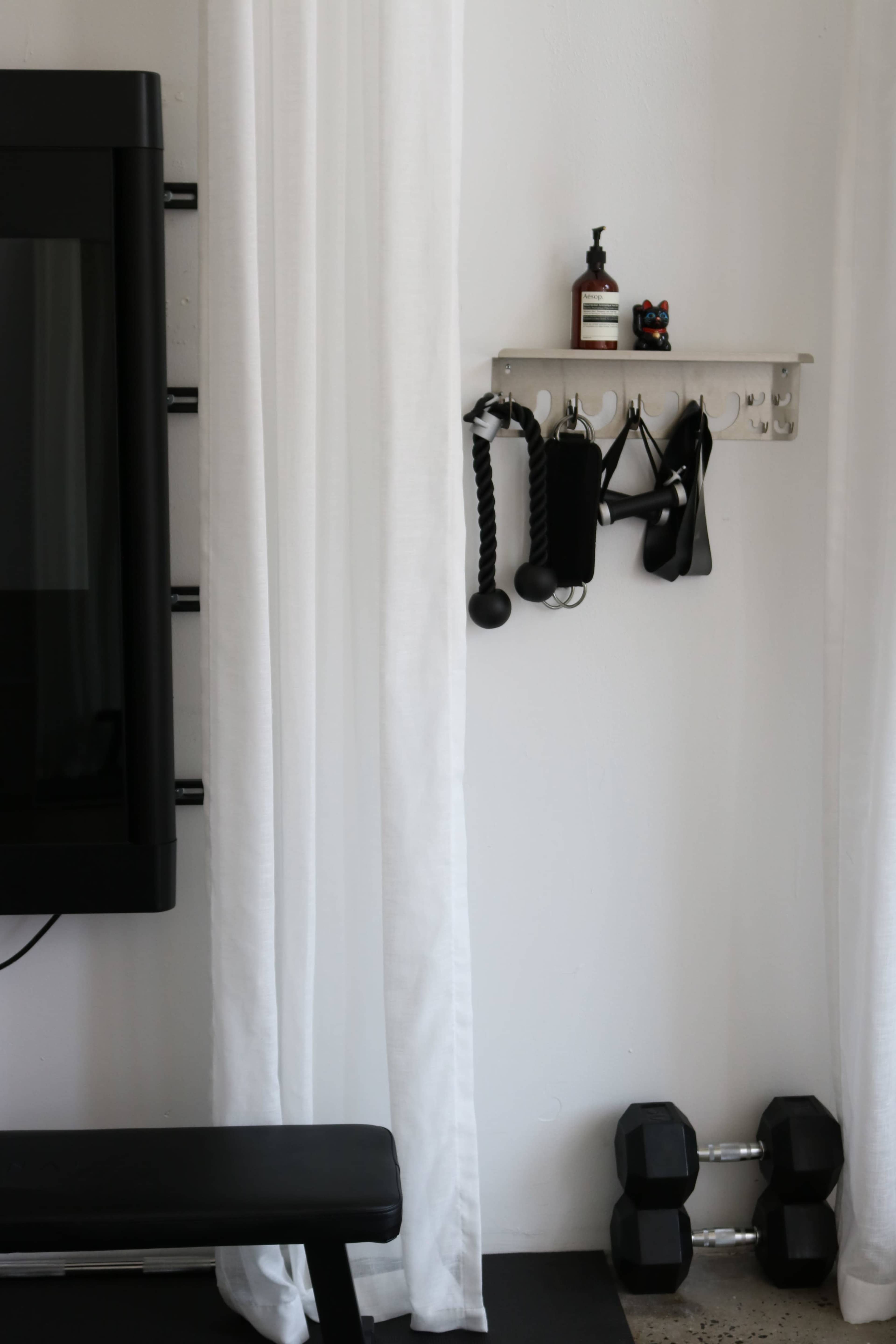 The image shows a small fitness area with a weight bench, a shelf holding fitness accessories, and a pair of dumbbells on the floor.