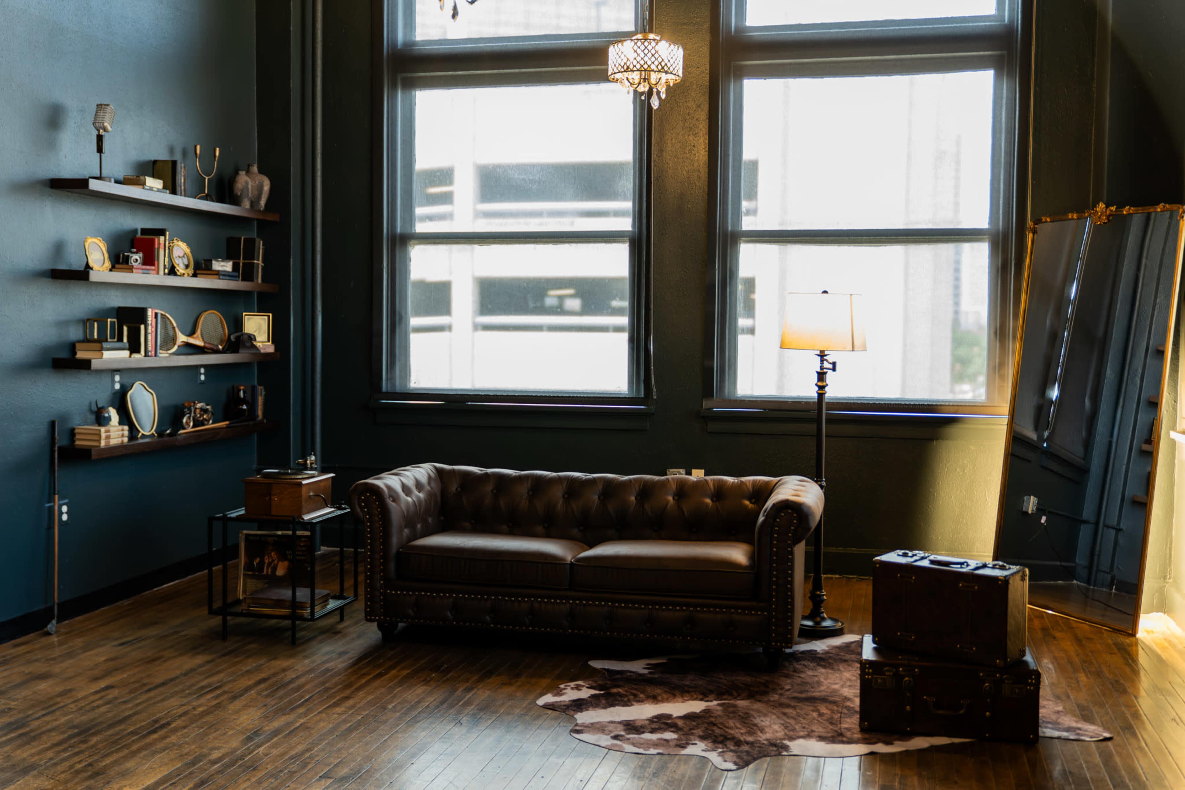 The image shows a cozy room with a brown leather couch, decorative shelves on the walls, a large mirror, a lamp, and two vintage suitcases on the floor.