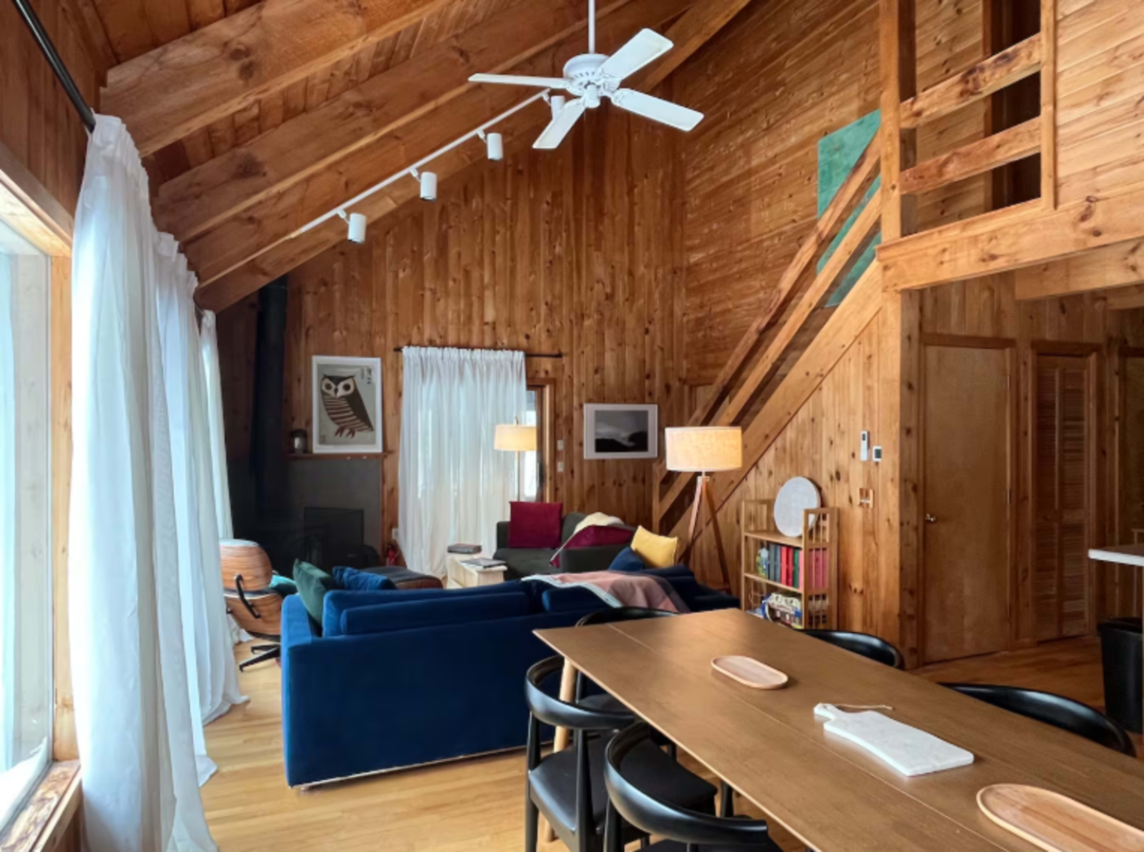 The image shows a cozy wooden cabin interior with a seating area featuring a blue sofa, a dining table, and an open staircase.