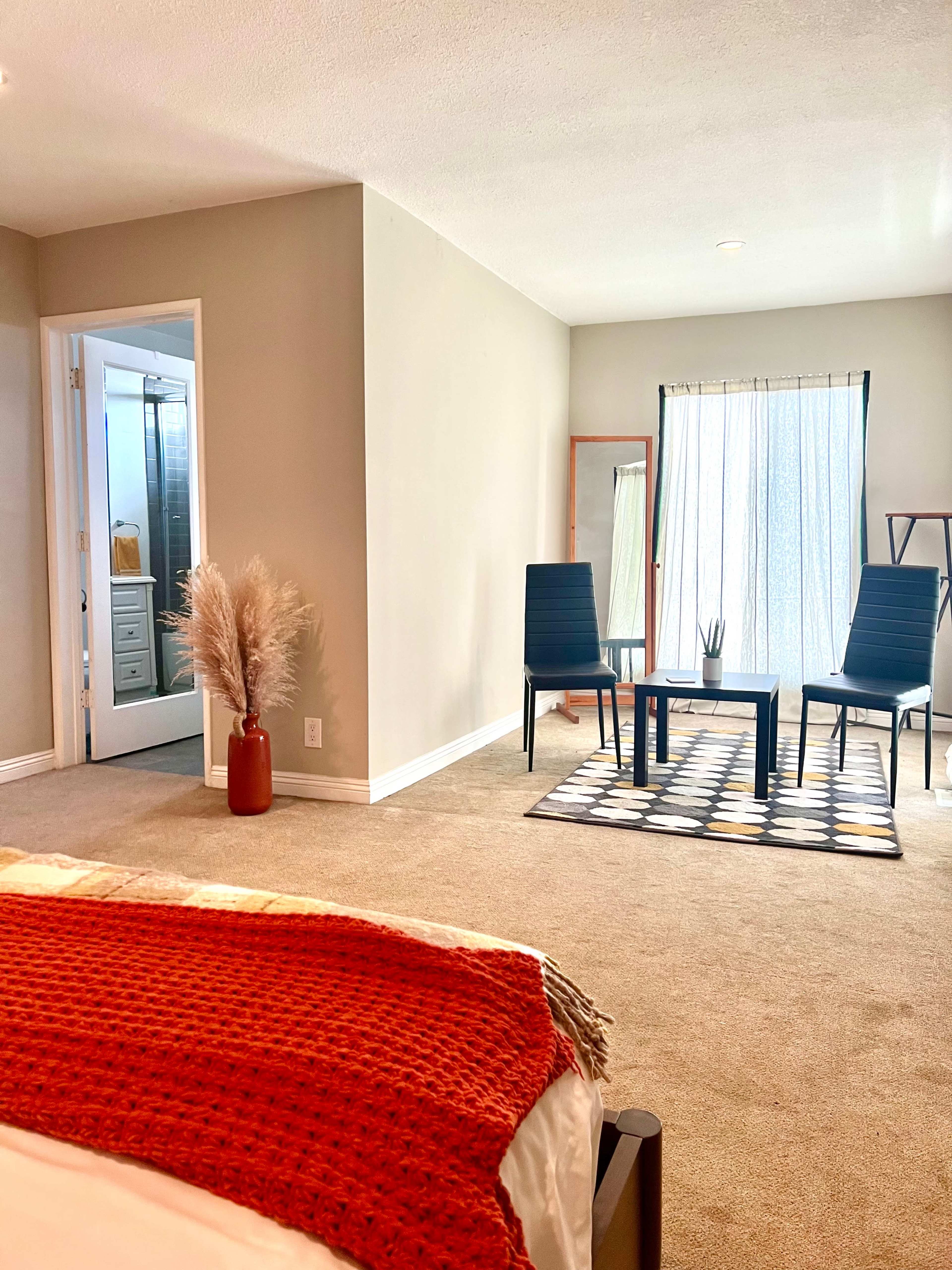 A spacious bedroom with two chairs and a table, featuring soft beige carpeting and large windows that allow natural light to enter.