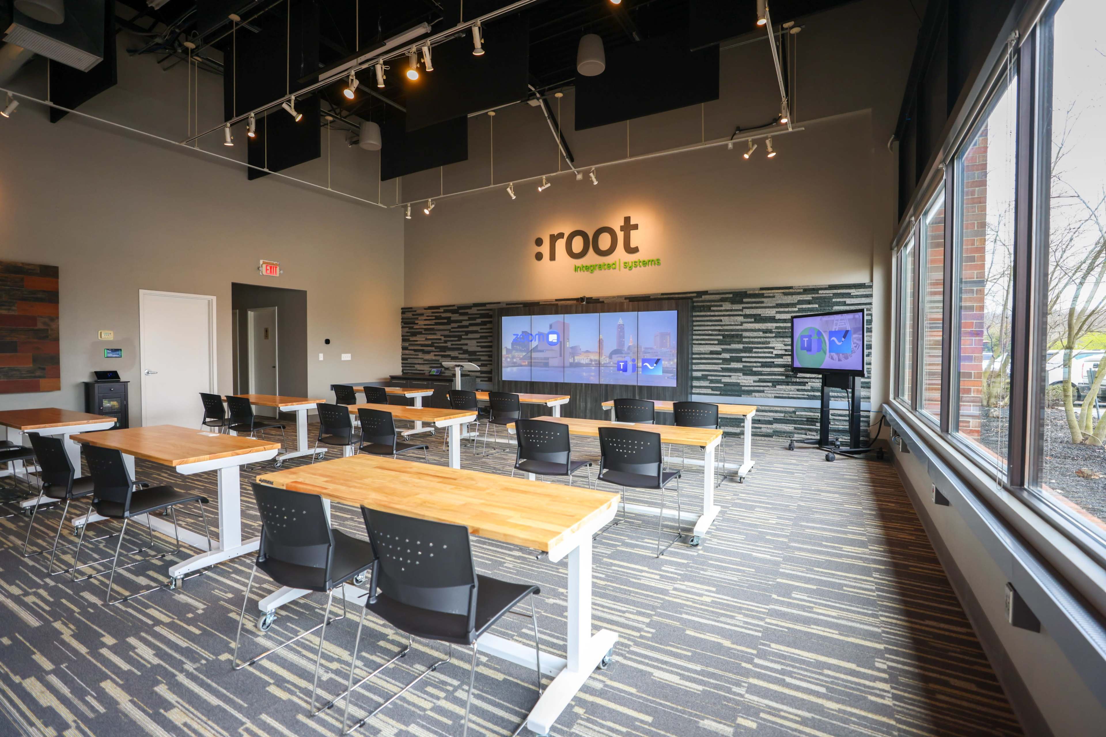 A modern classroom with multiple wooden tables and black chairs arranged facing a large screen on the wall.