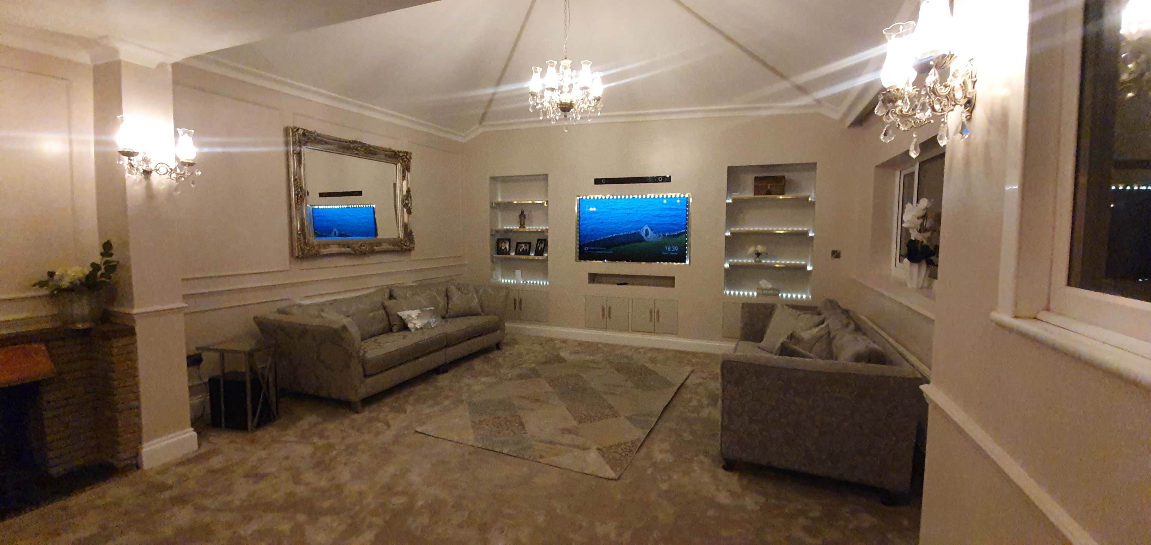 The room features a living area with two sofas, a wall-mounted television, decorative shelves, and chandeliers.