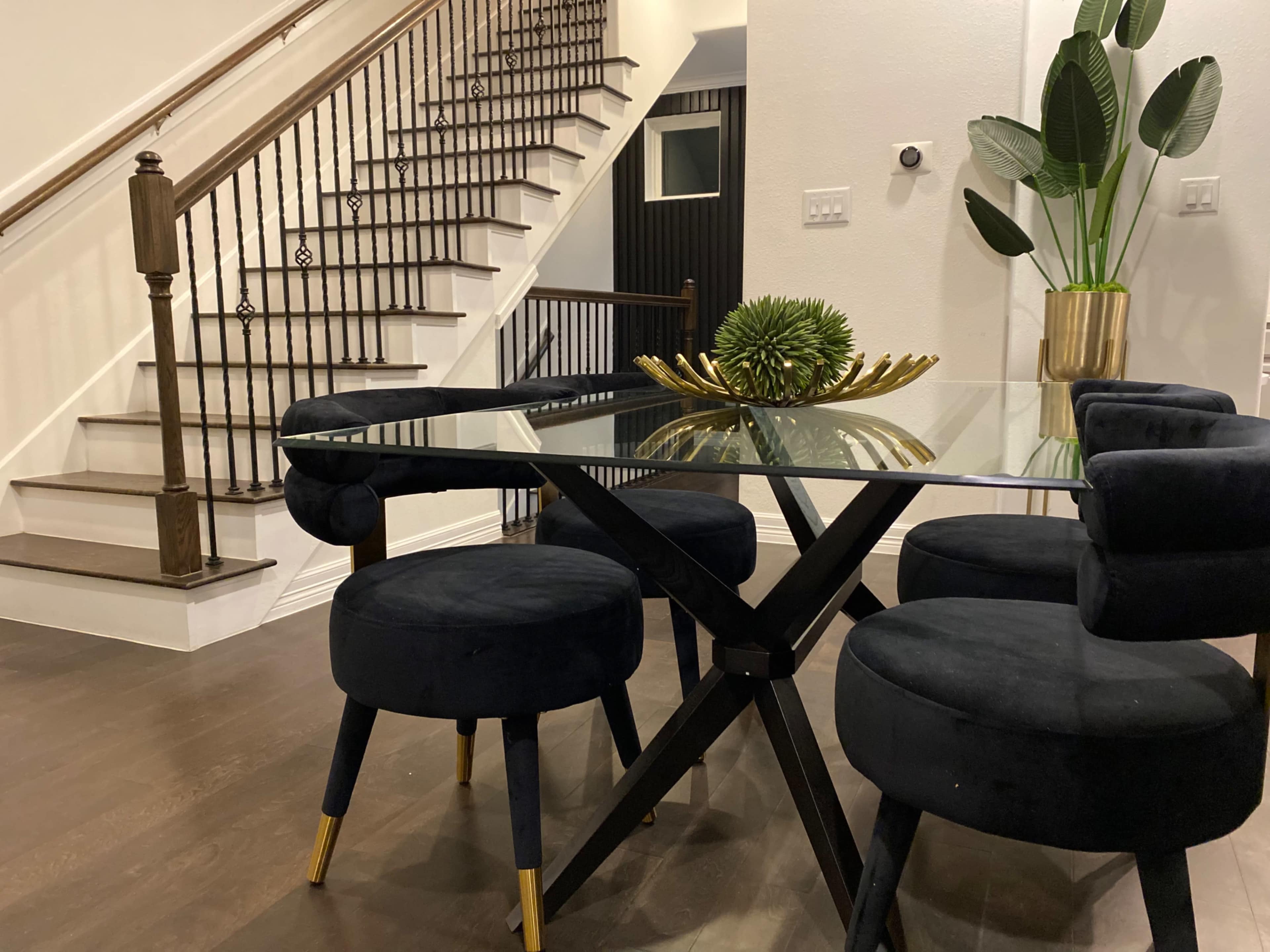 A modern dining area features a glass-top table surrounded by four navy blue upholstered chairs, with a staircase and potted plant in the background.