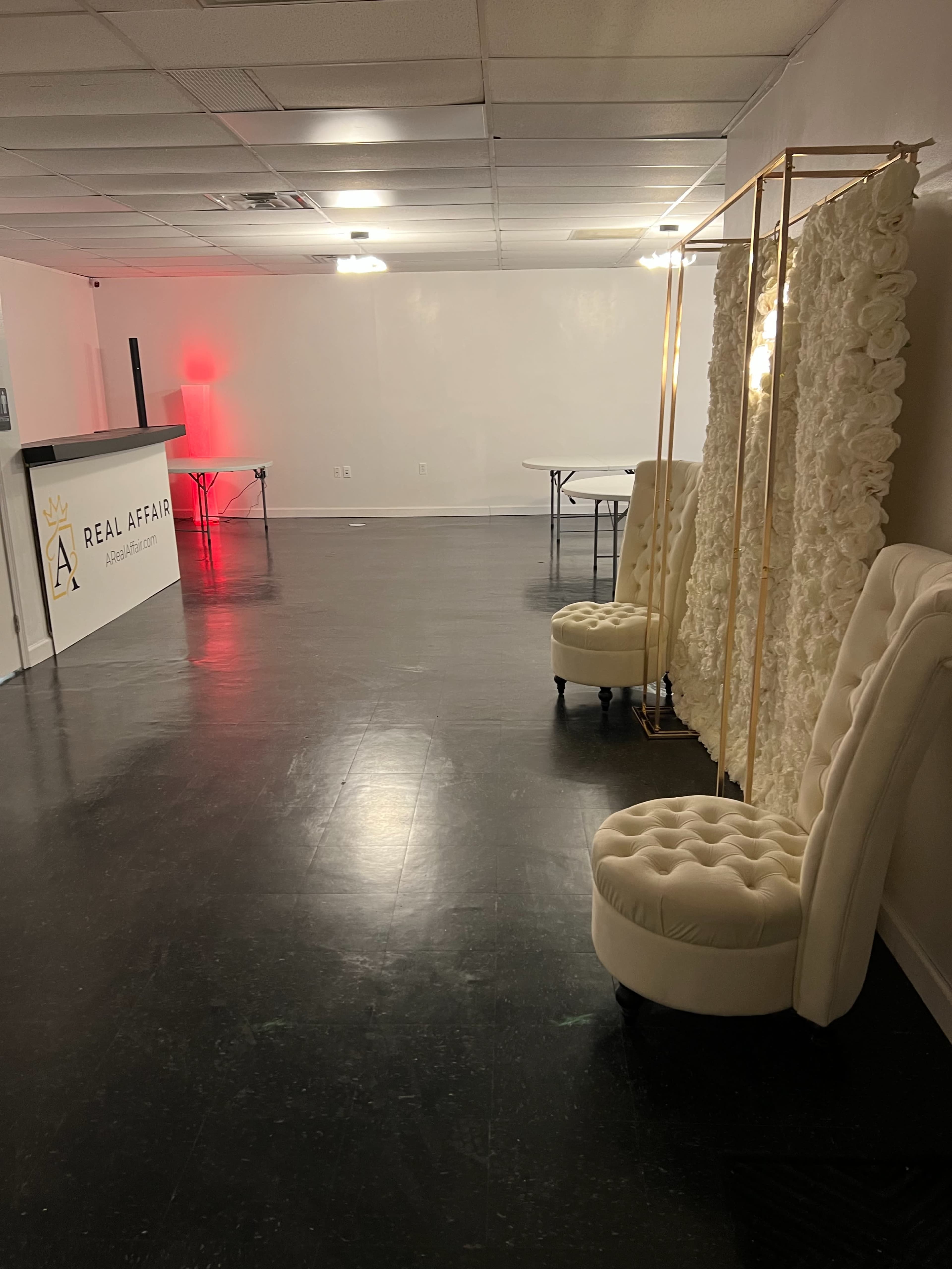 A spacious, empty room with a white wall, a reception desk labeled "A REAL AFFAIR," two plush white chairs, and decorative floral panels set up against one side.