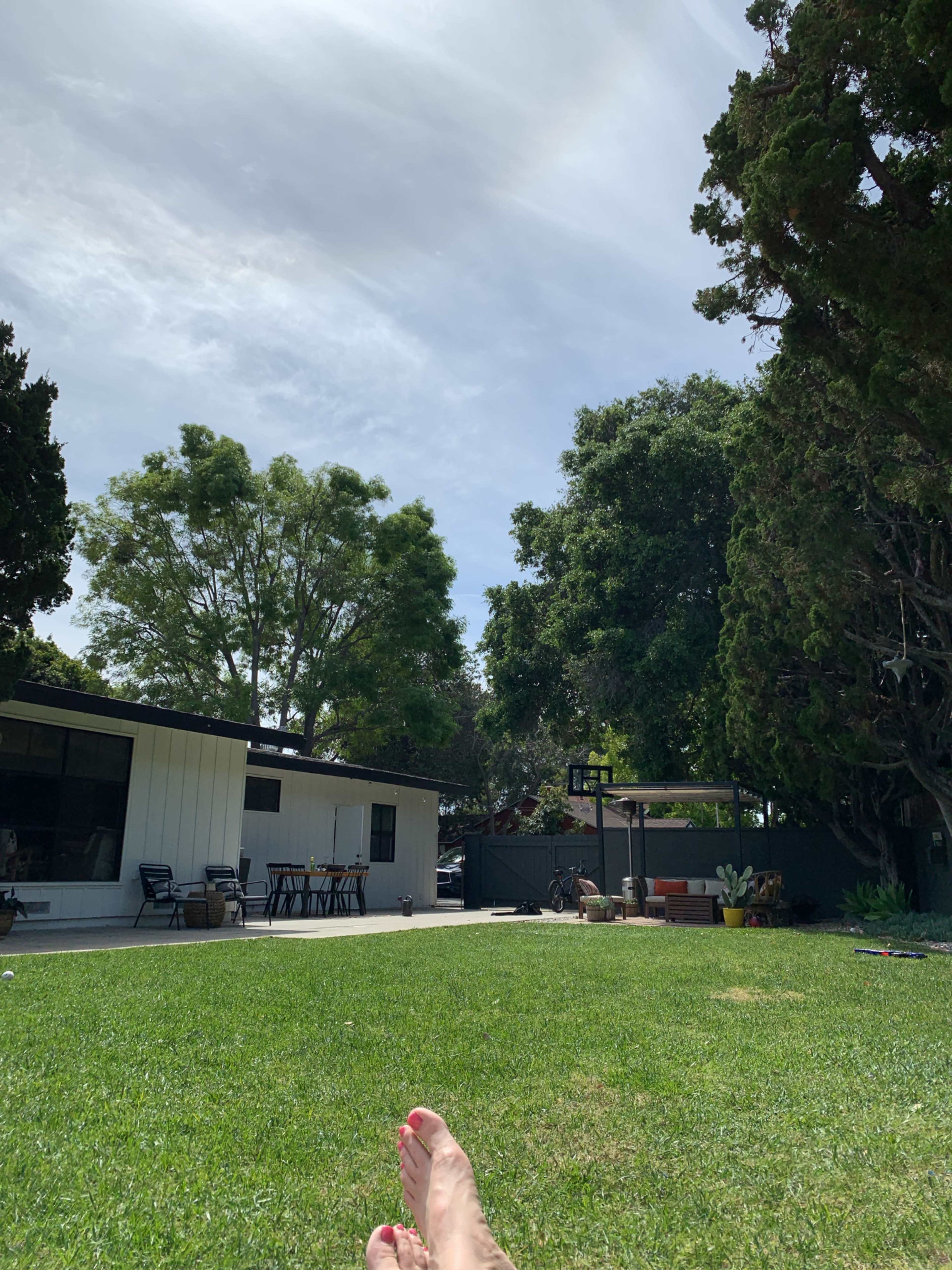 A person is relaxing on a green lawn in a backyard with trees and a house in the background under a partly cloudy sky.